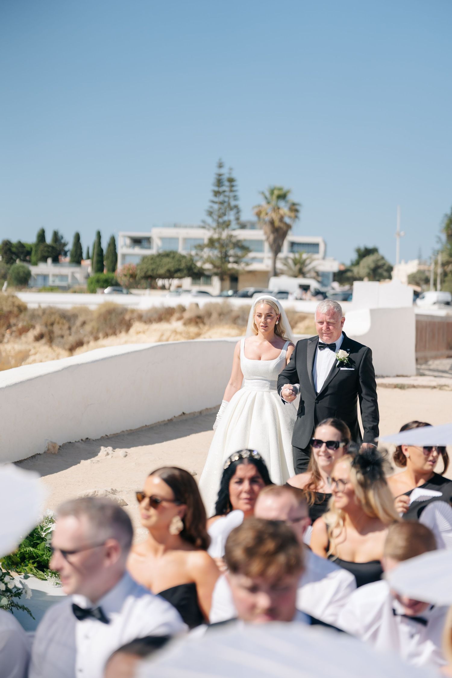 Wedding ceremony guests in white attire gather outdoors under bright blue skies.