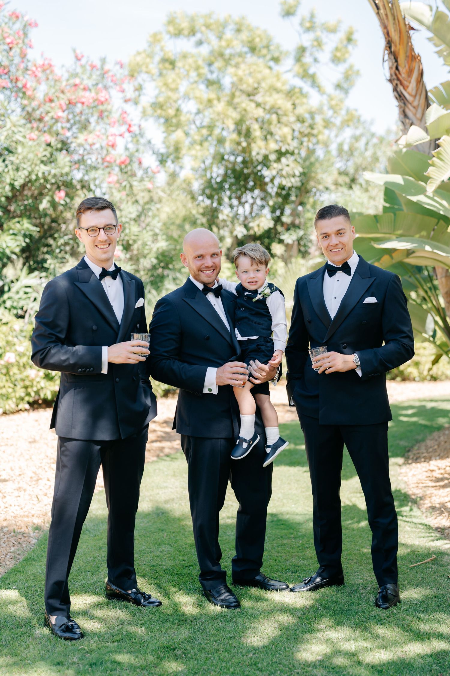Wedding party in tuxedos enjoying drinks and laughs outdoors.