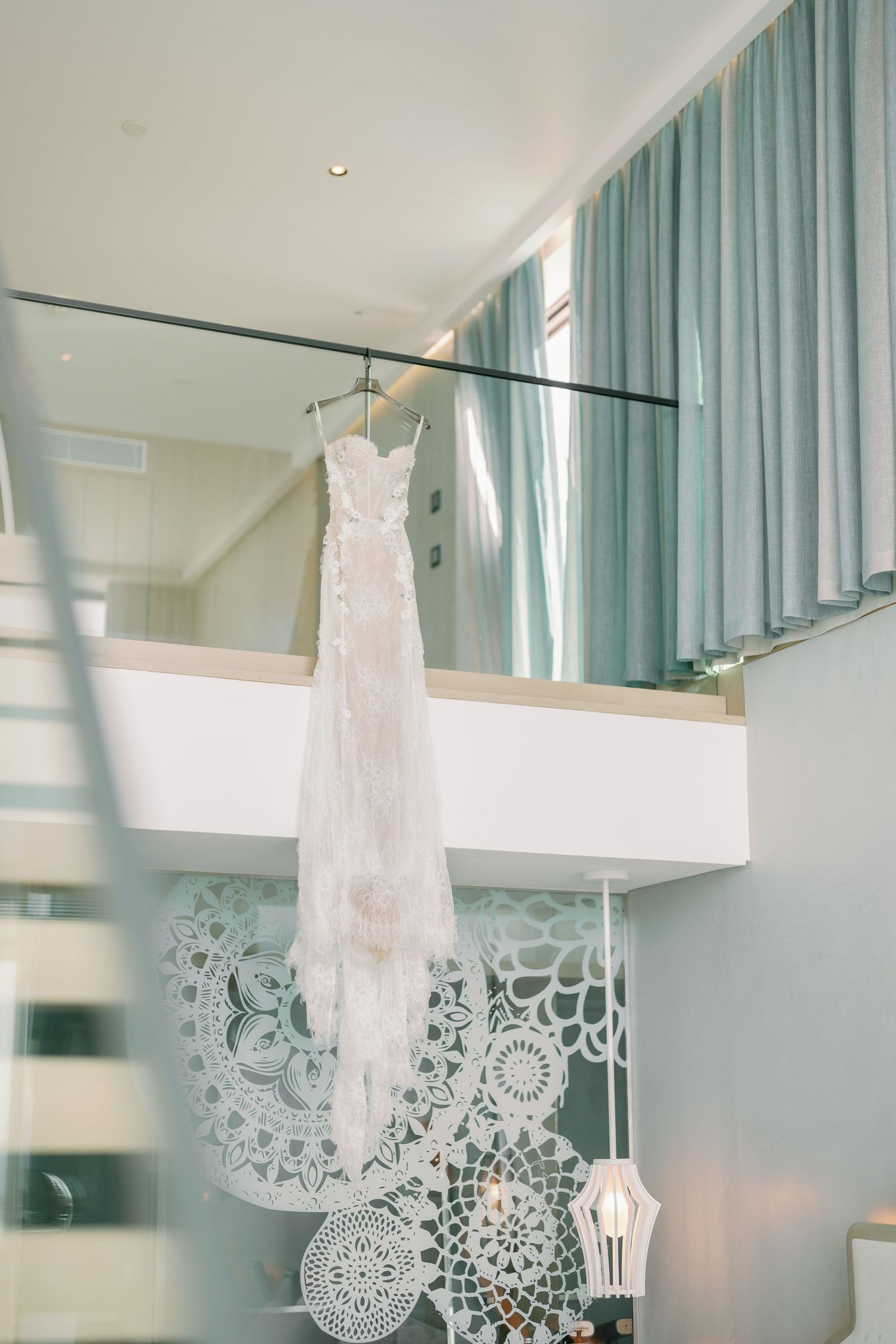Elegant white lace wedding dress hanging in modern room with decorative backdrop.