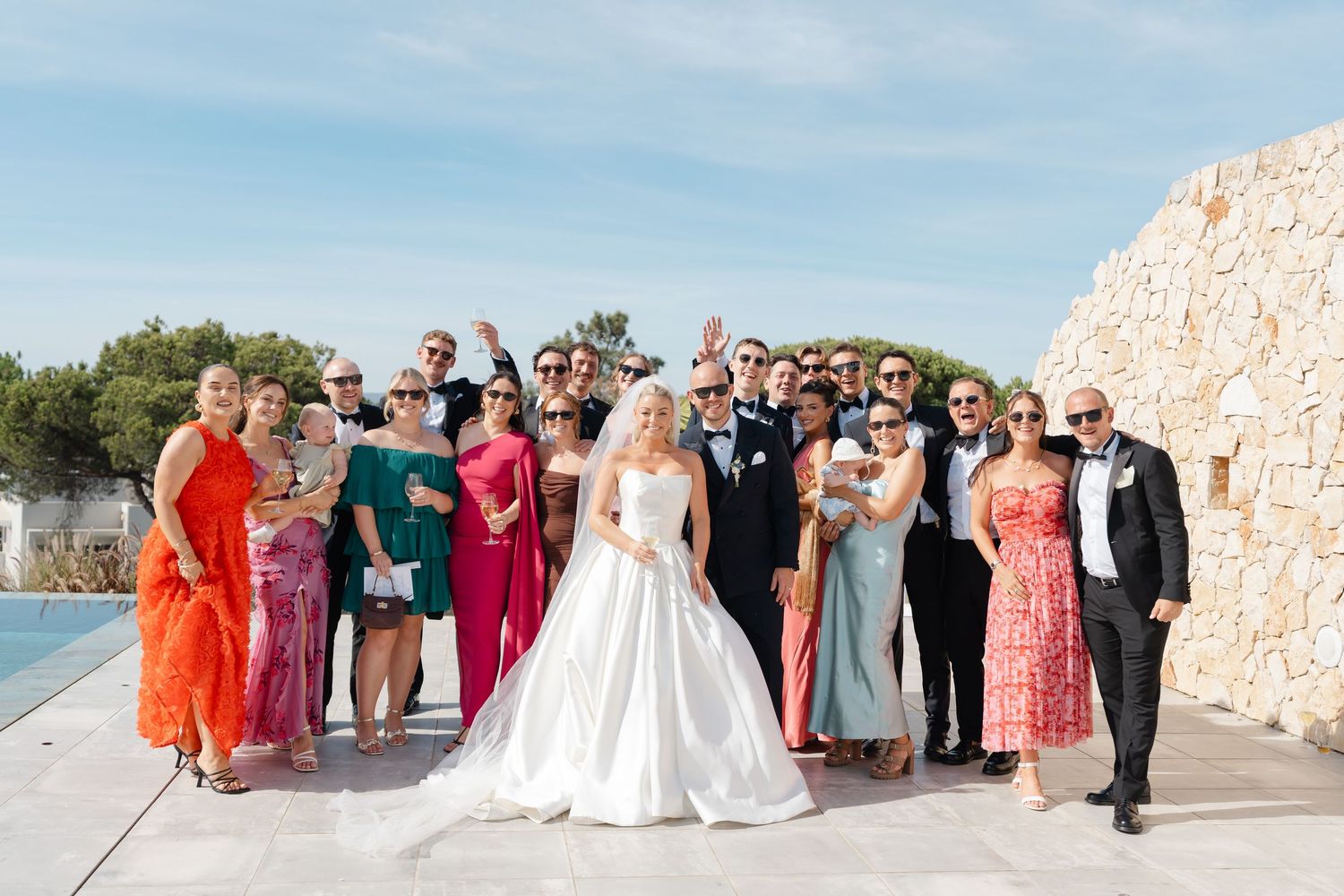 Wedding party and guests gathered for group photo at scenic waterfront location.