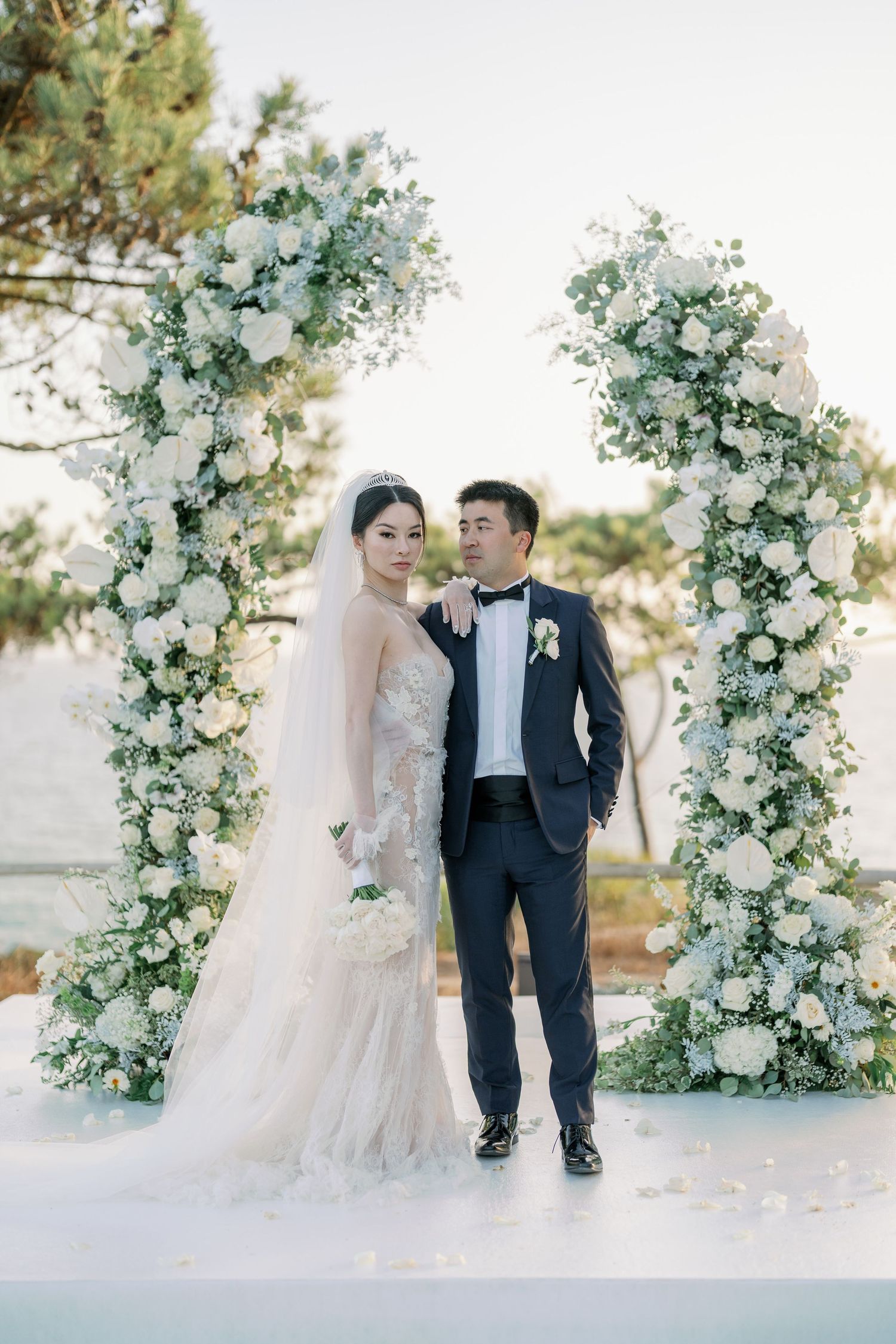 Couple poses under an elegant white floral arch at their outdoor wedding ceremony.