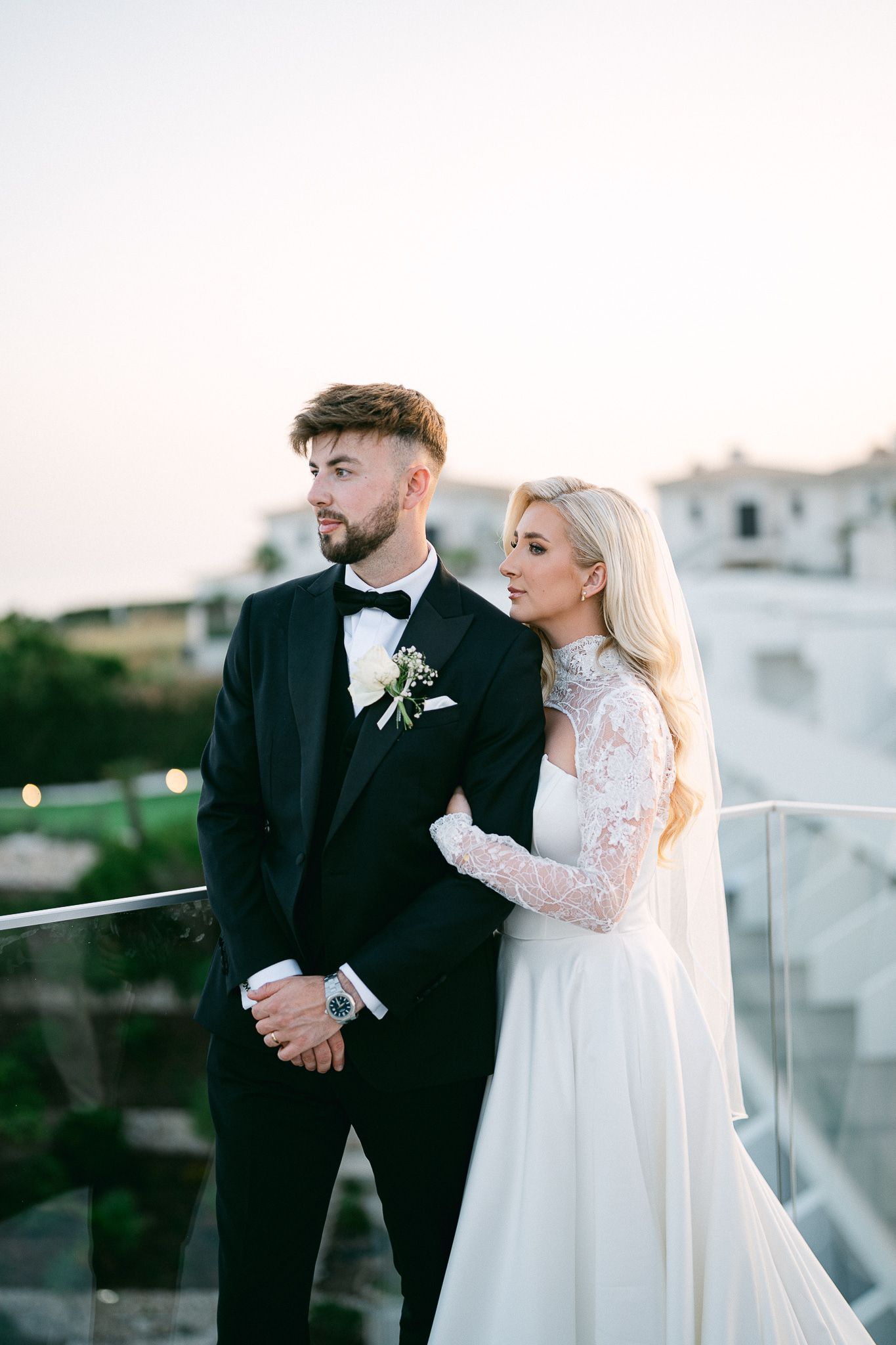 Elegant bride in lace sleeves and groom in tuxedo share romantic moments during outdoor photo session.
