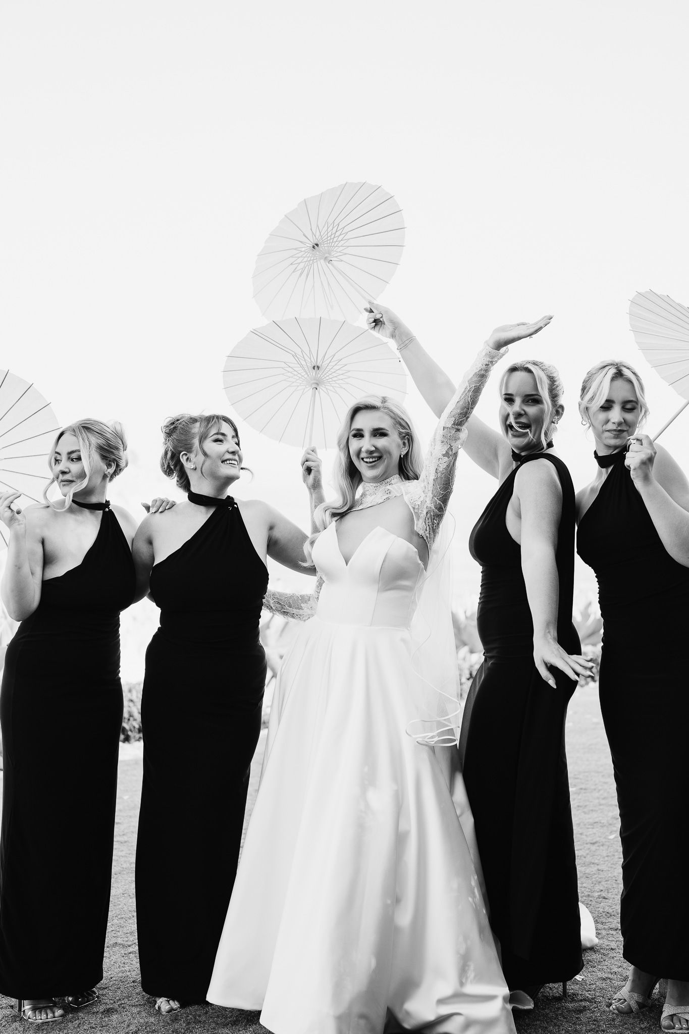 Bridal party in black dresses poses with white parasols alongside bride in elegant white gown.
