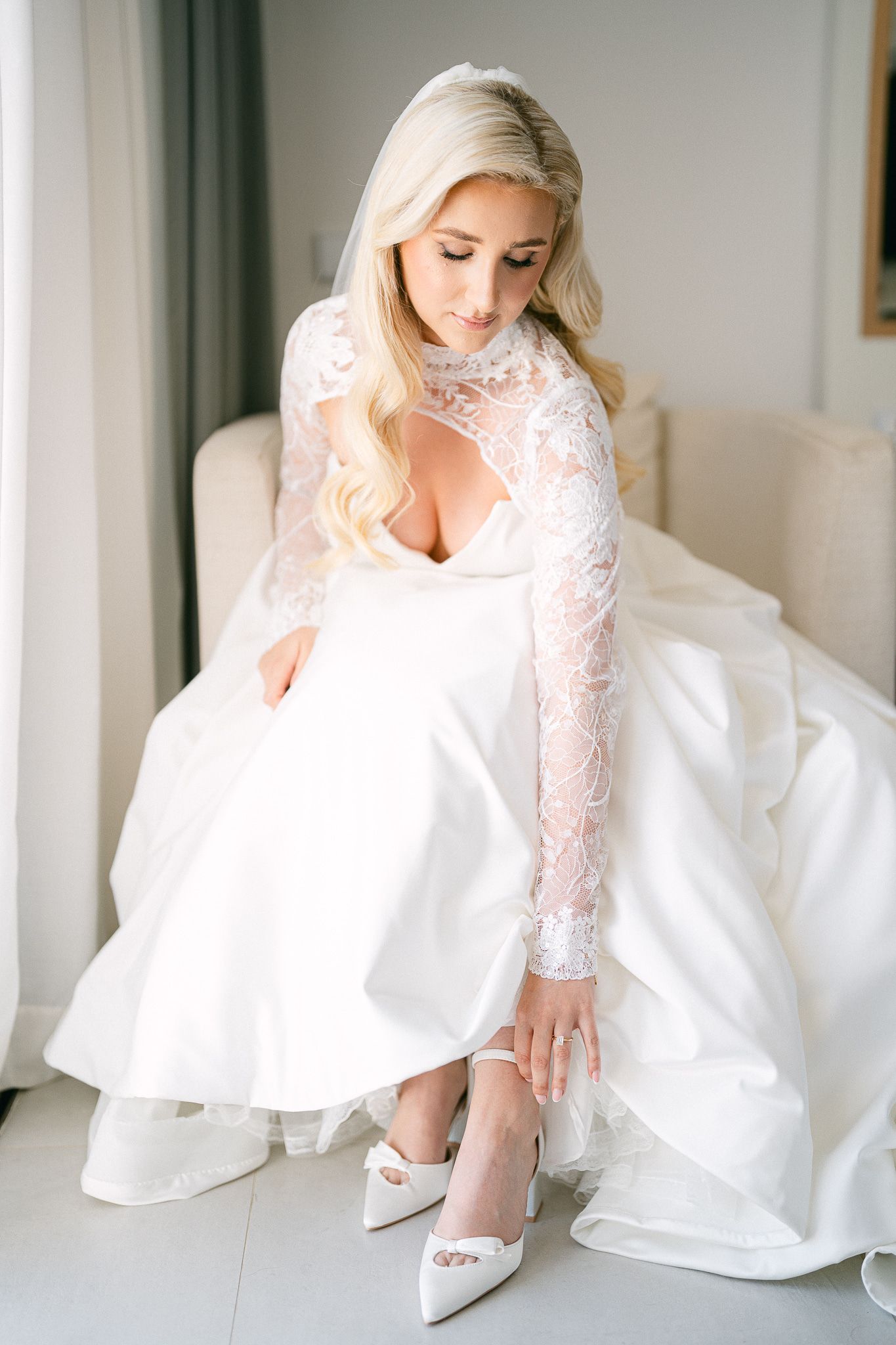 A bride in a white lace gown adjusts her shoe in an elegant indoor setting.