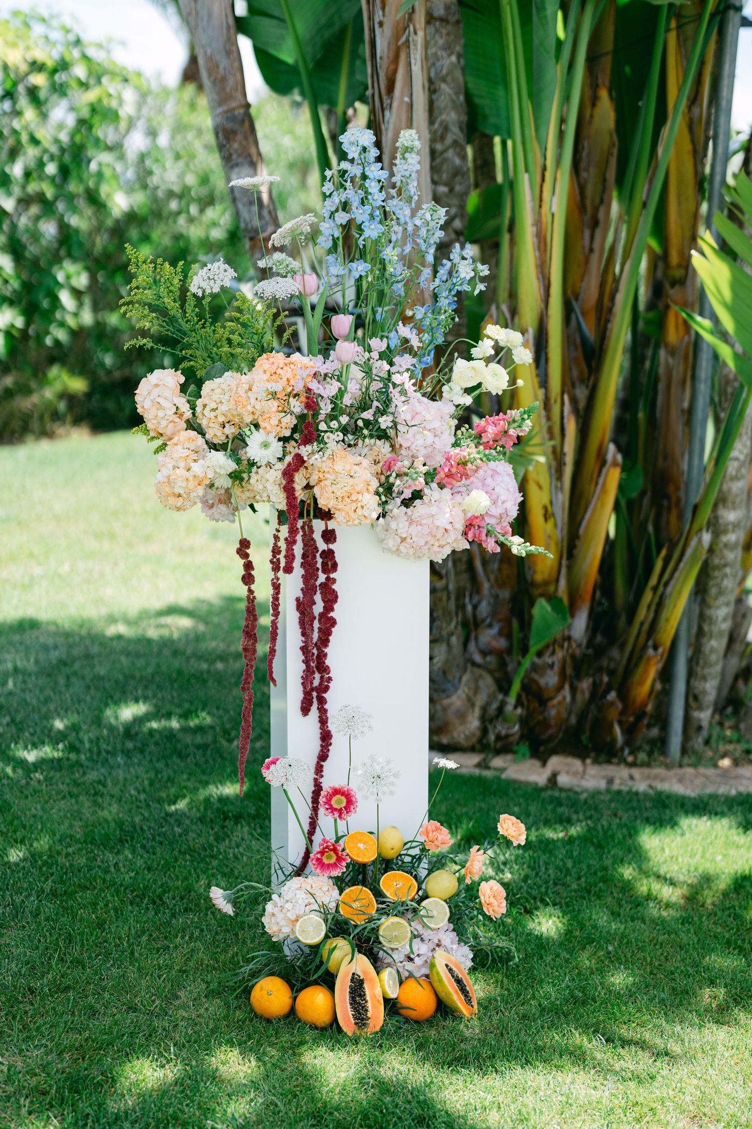A stunning floral arrangement with citrus fruits and pink blooms on a white pedestal outdoors.