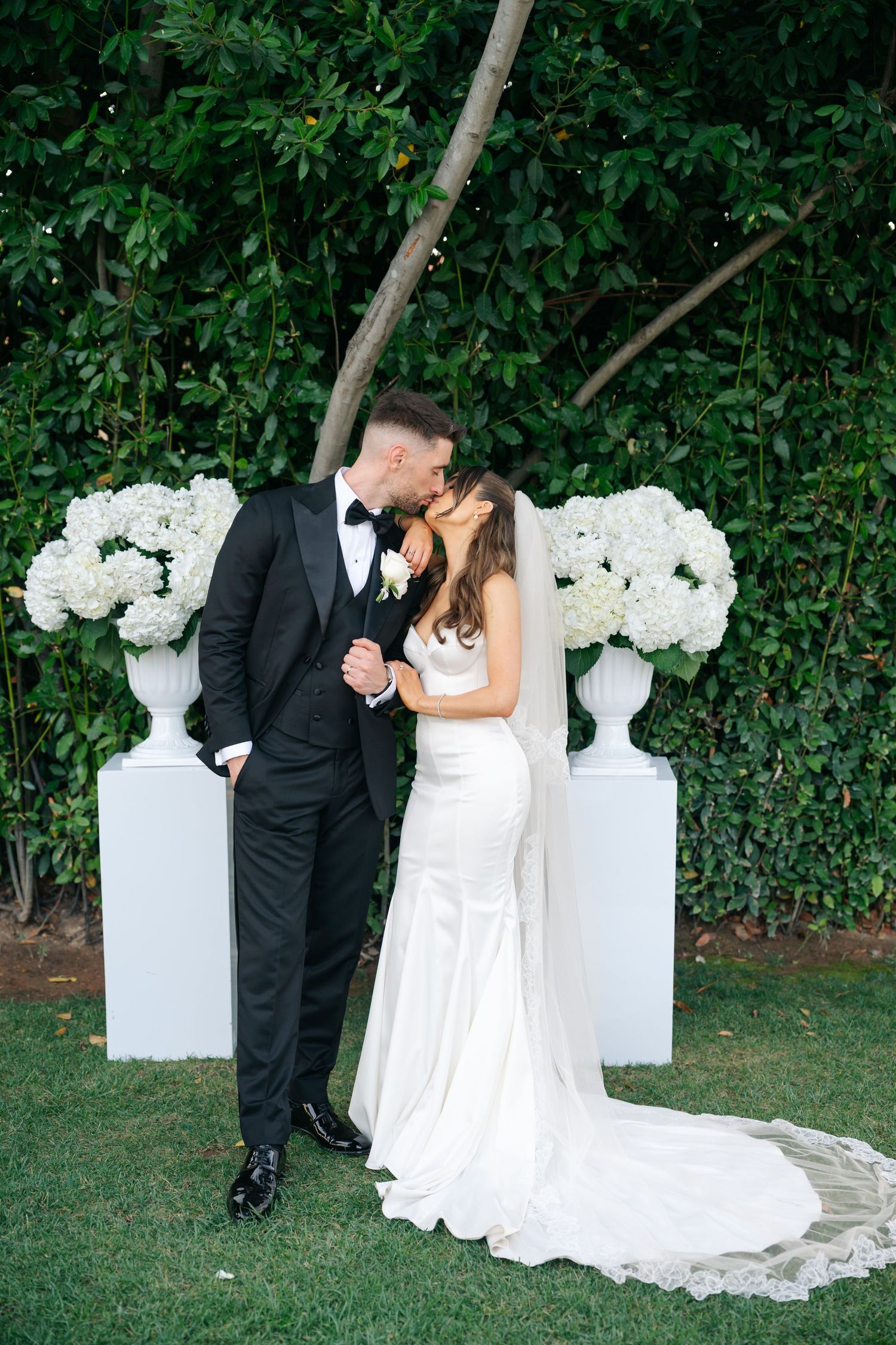 Wedding couple poses against greenery backdrop with elegant white floral arrangements.