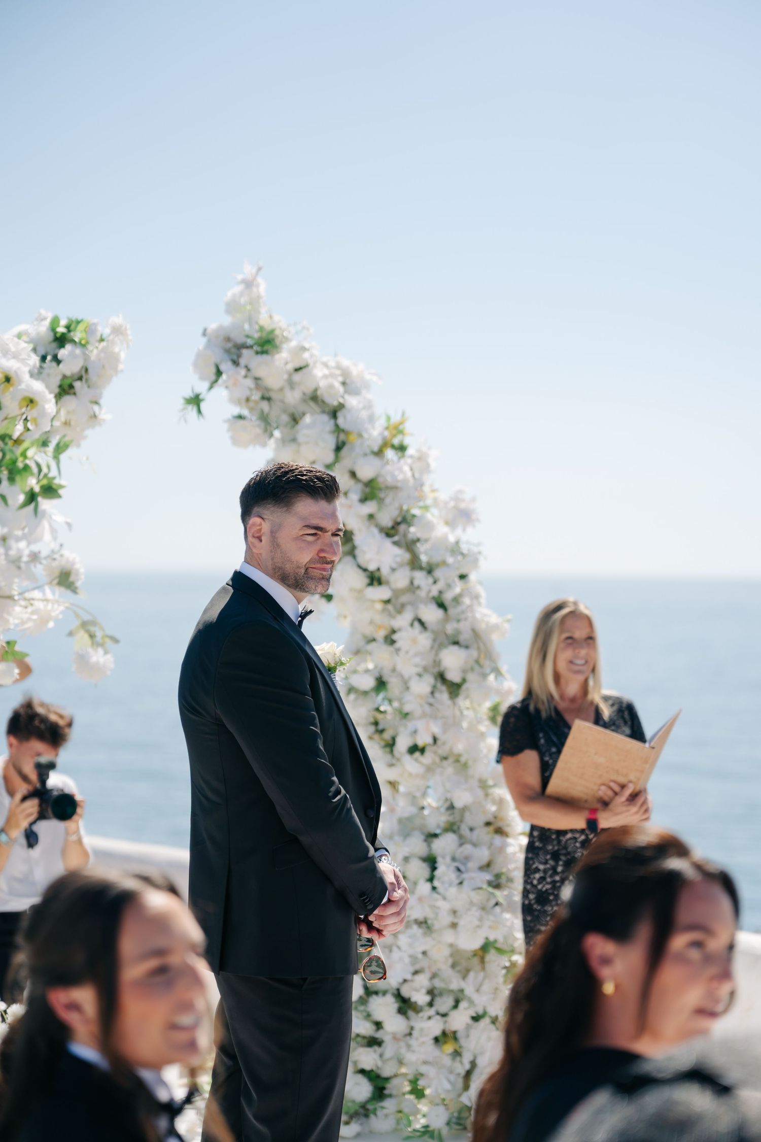 Elegant wedding ceremony setup with white floral arches against an ocean backdrop.