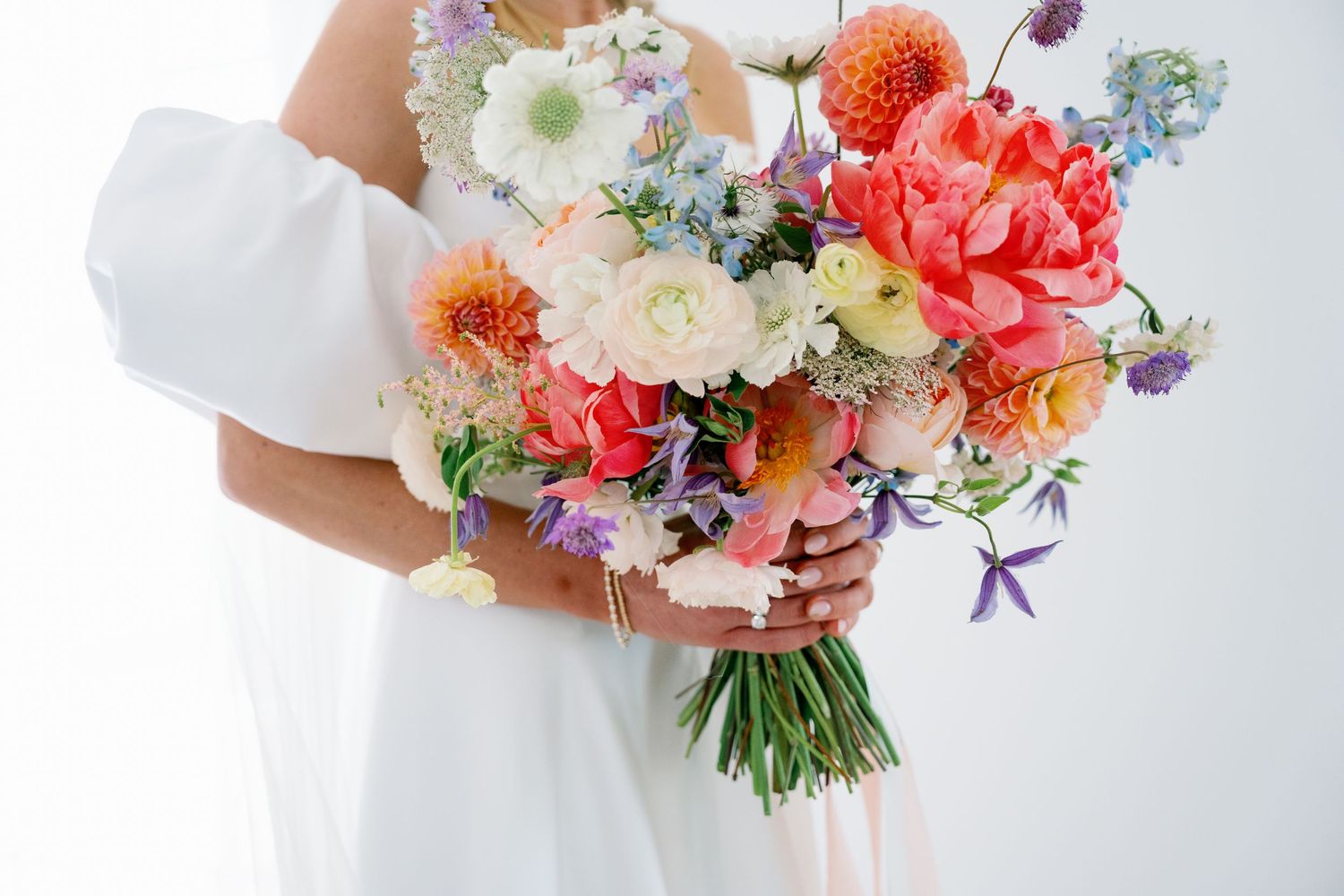 A colorful wedding bouquet featuring coral dahlias, white daisies, and purple delphinium flowers held by someone in white.