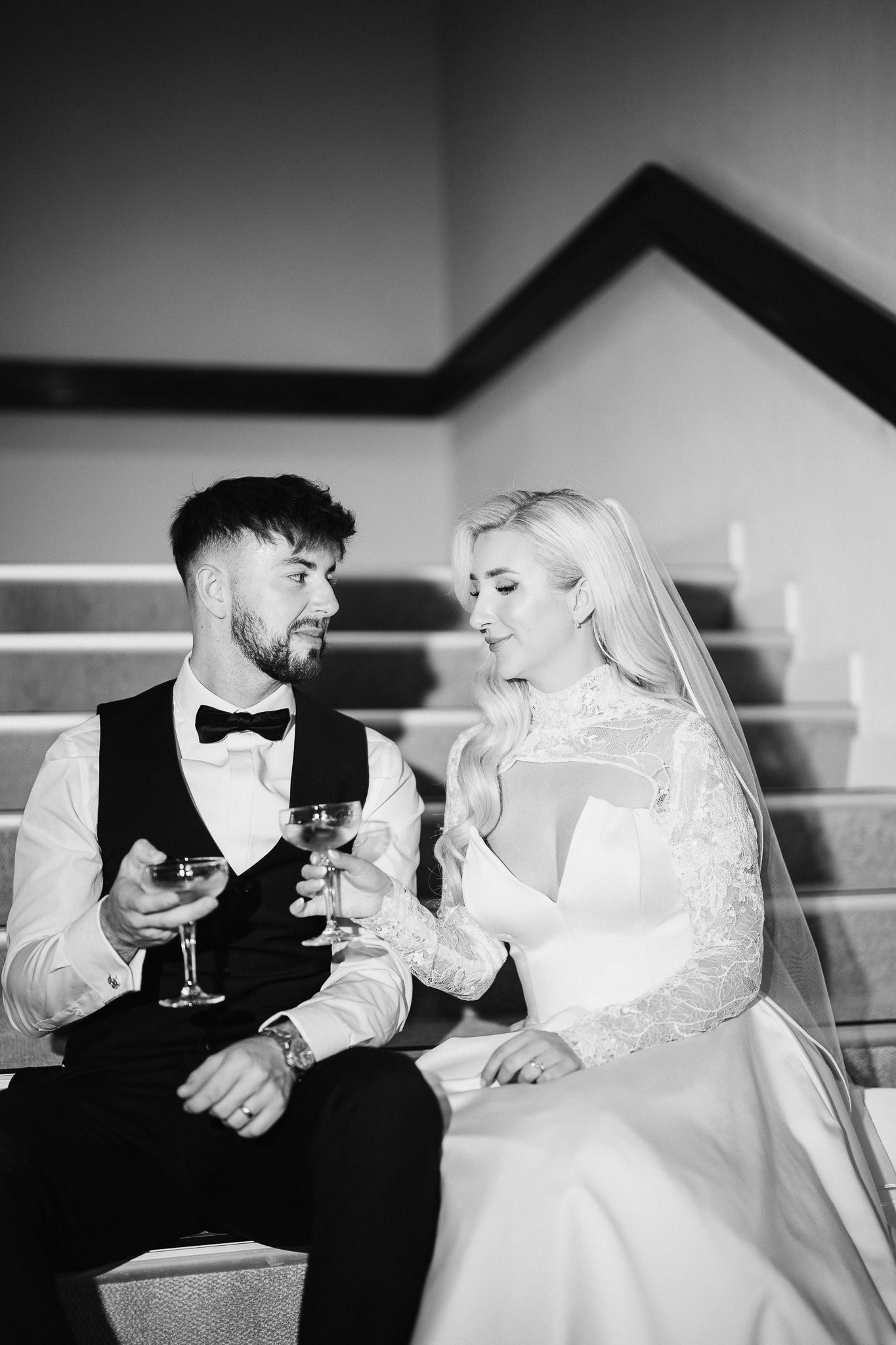 A bride in white and groom in black sit together sharing drinks at their wedding.