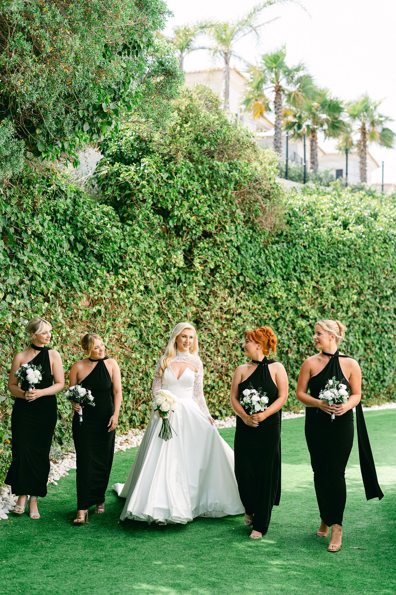 Wedding party in black dresses and white gown poses in front of lush green hedges outdoors.