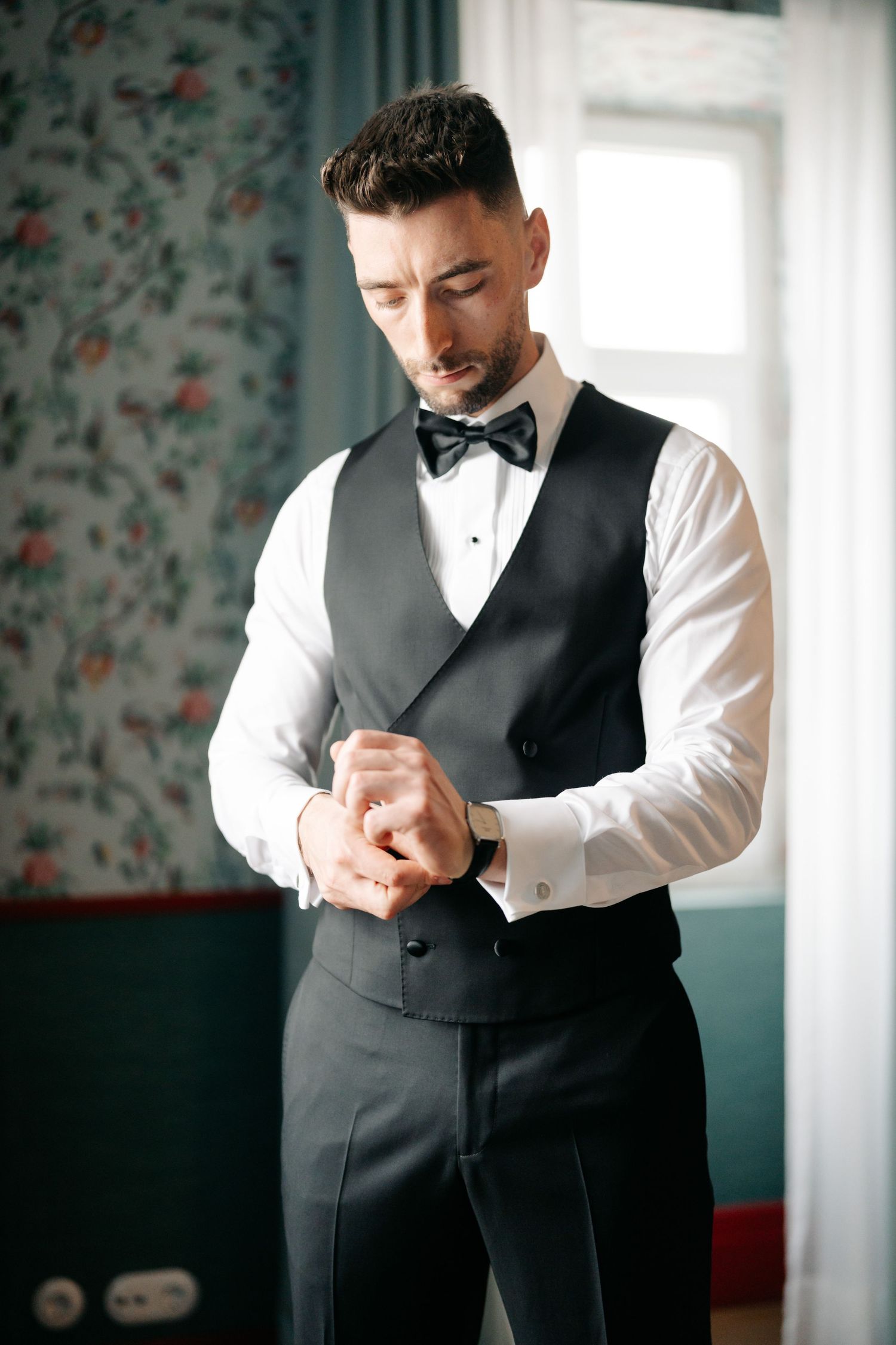Formal tuxedo attire with black bow tie and vest being worn in an elegant room with floral wallpaper.