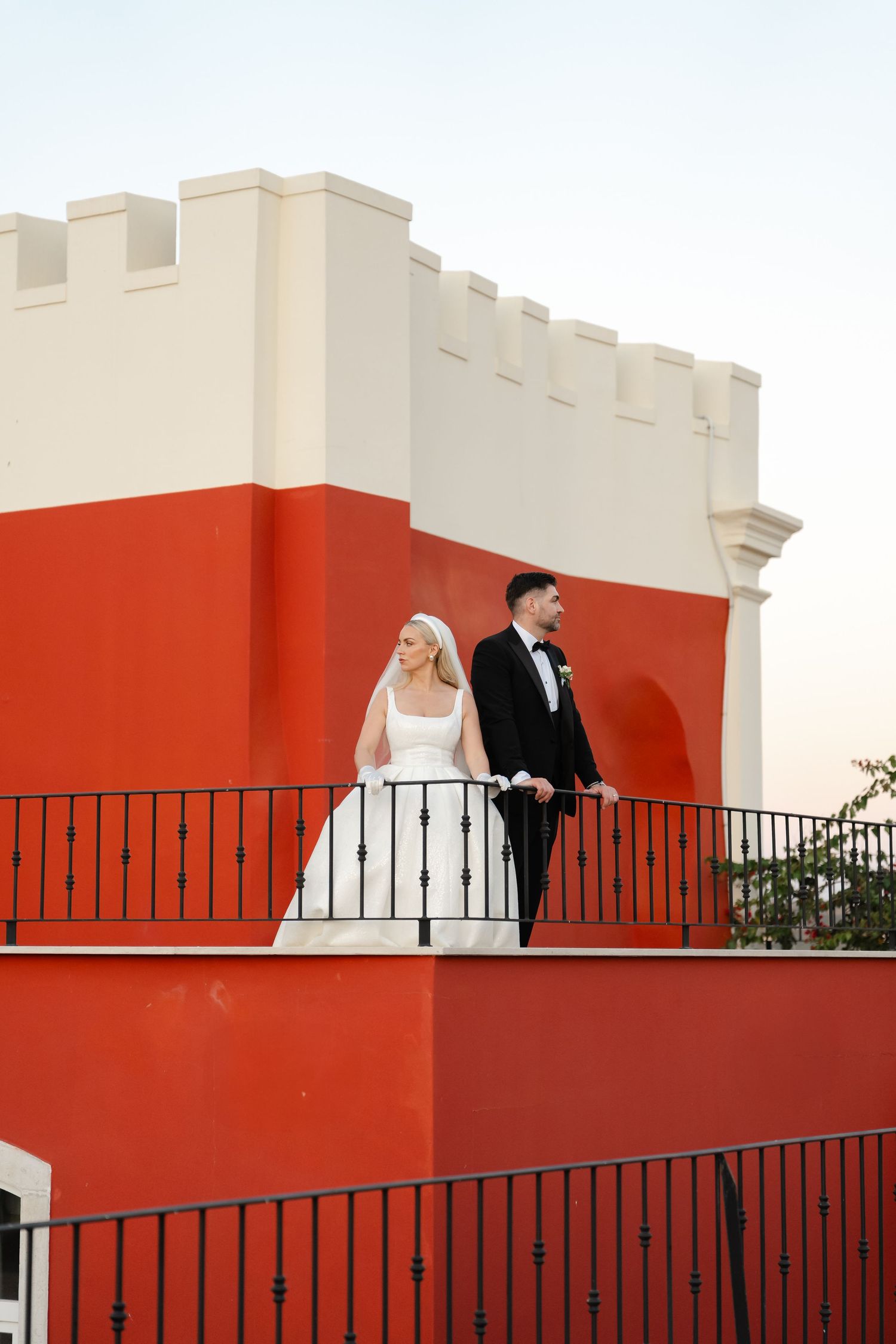 Wedding couple stands on a red and white architectural balcony against clear sky.