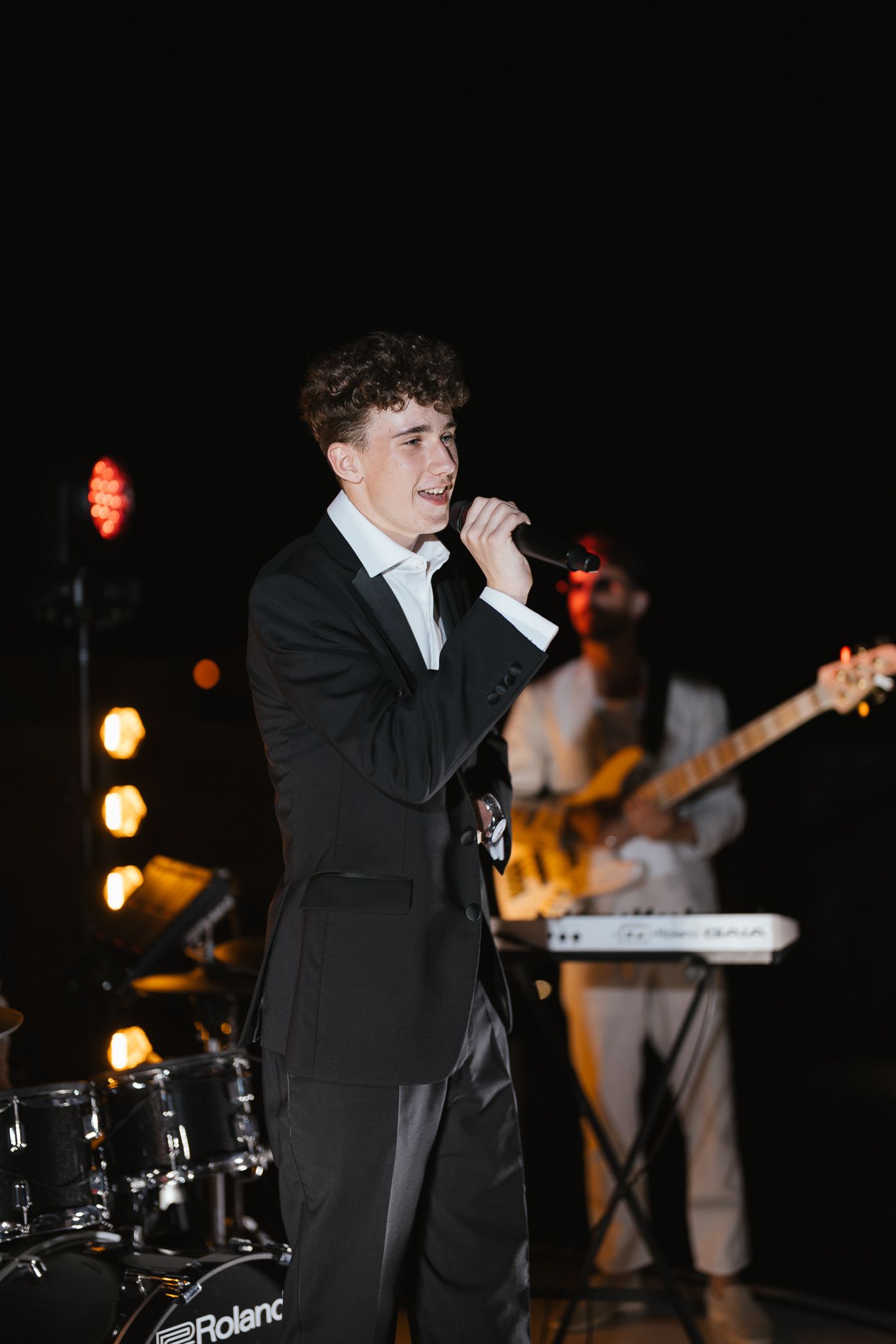 Singer in formal attire performing with band at nighttime event.