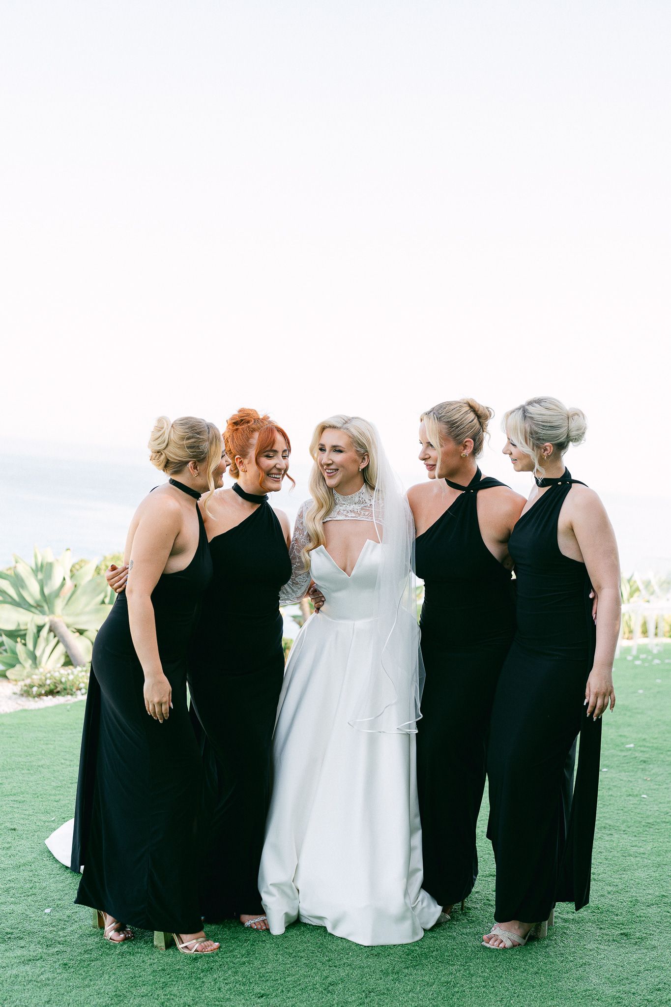 Bridal party in coordinated black dresses stands with bride in white gown on green lawn.