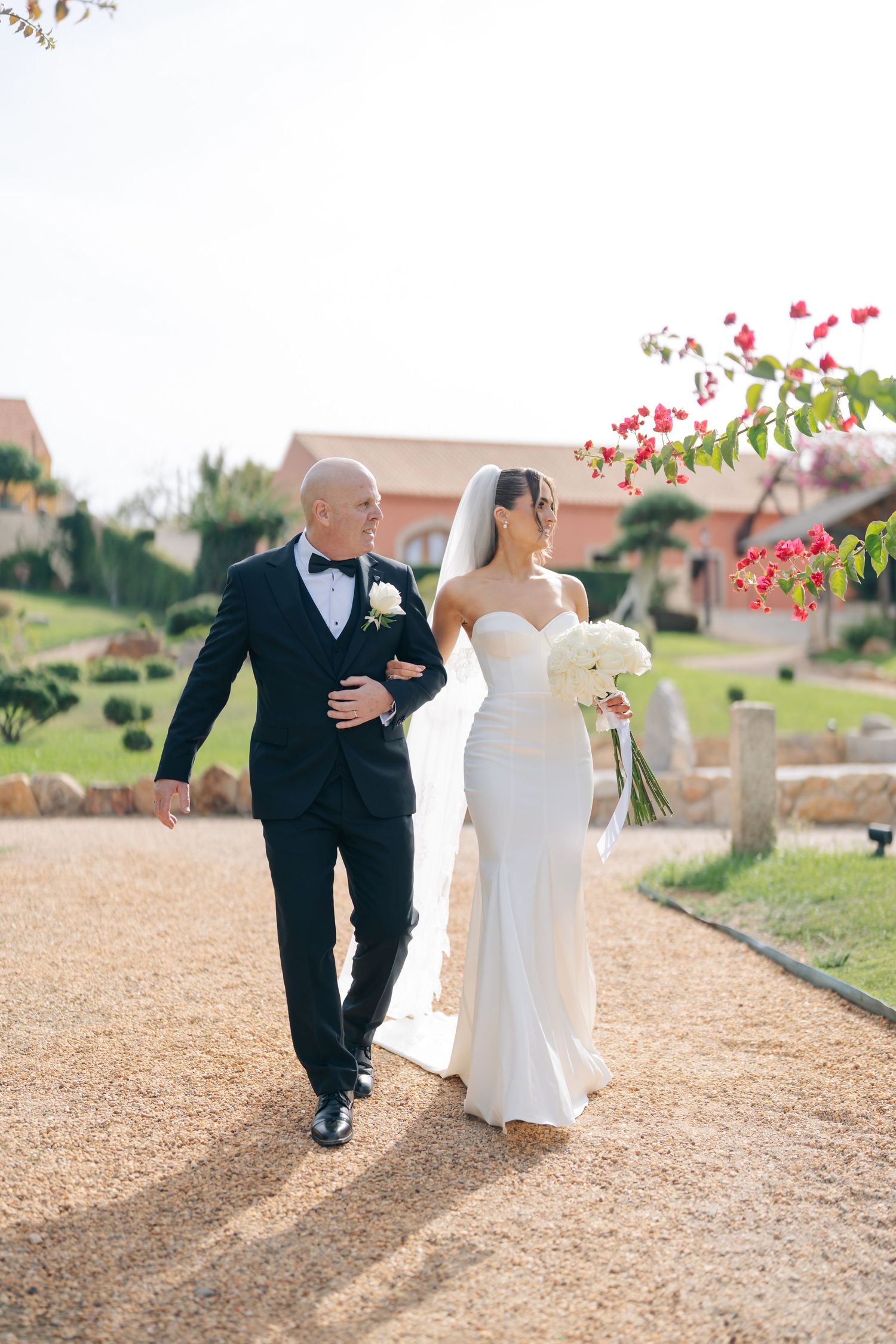 A couple walks along a sunlit path with pink bougainvillea flowers in the background.