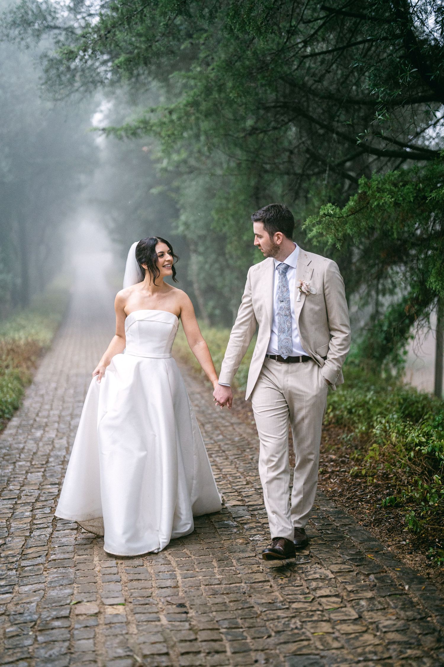 A bride and groom walk together on a misty cobblestone path surrounded by green trees and fog.