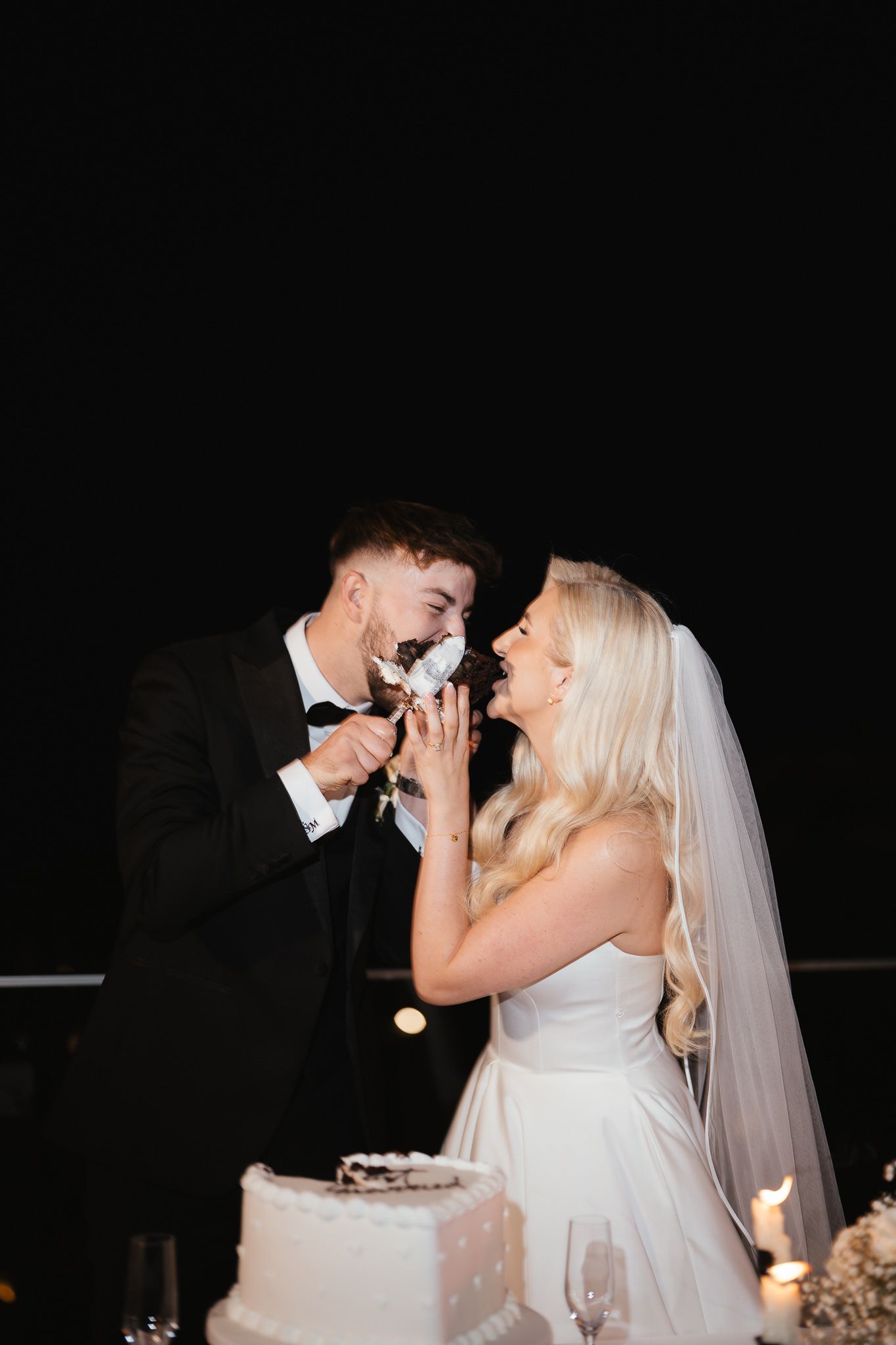Bride and groom share messy cake-feeding moment at wedding reception.