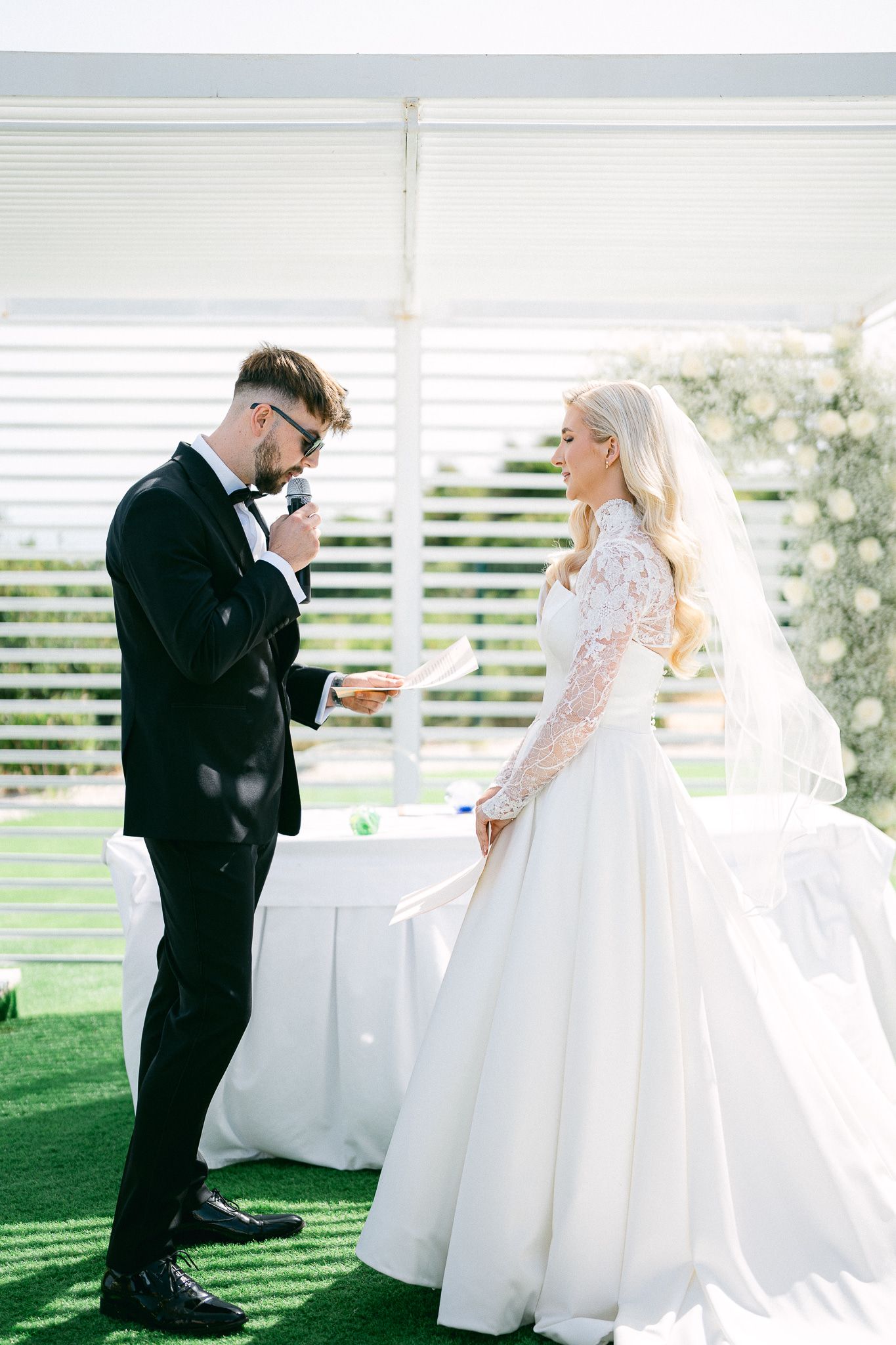 Wedding couple shares intimate first look moment under white pergola on manicured lawn.