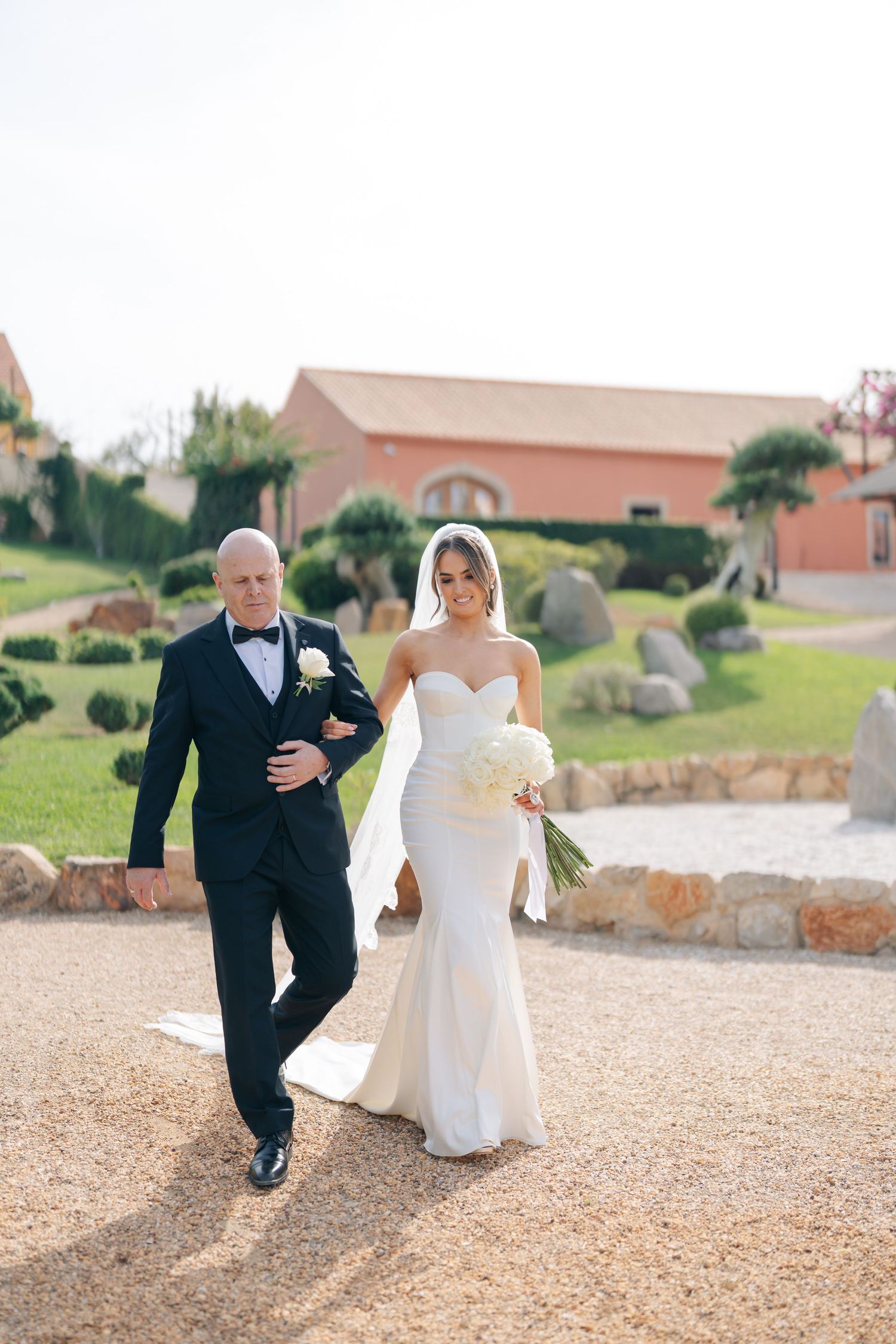 Wedding party members walk along a gravel path outside a venue.