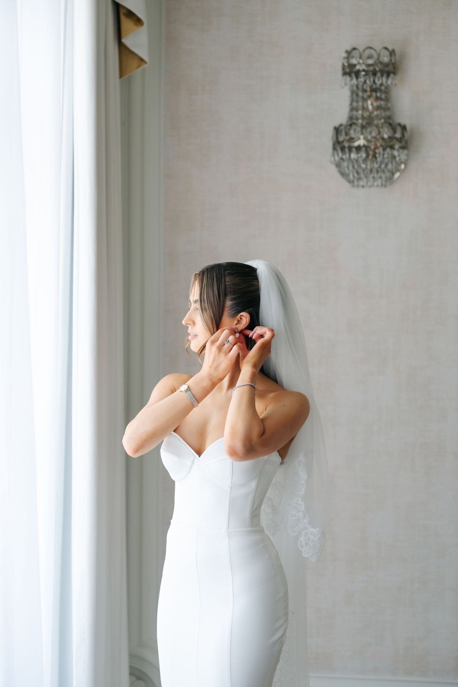 A bride in a white wedding dress poses playfully in an elegant room with a crystal chandelier.