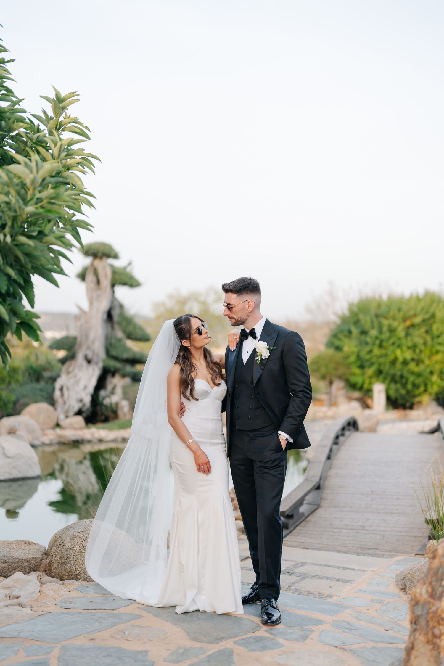 Wedding couple embraces on stone pathway surrounded by lush garden landscaping.