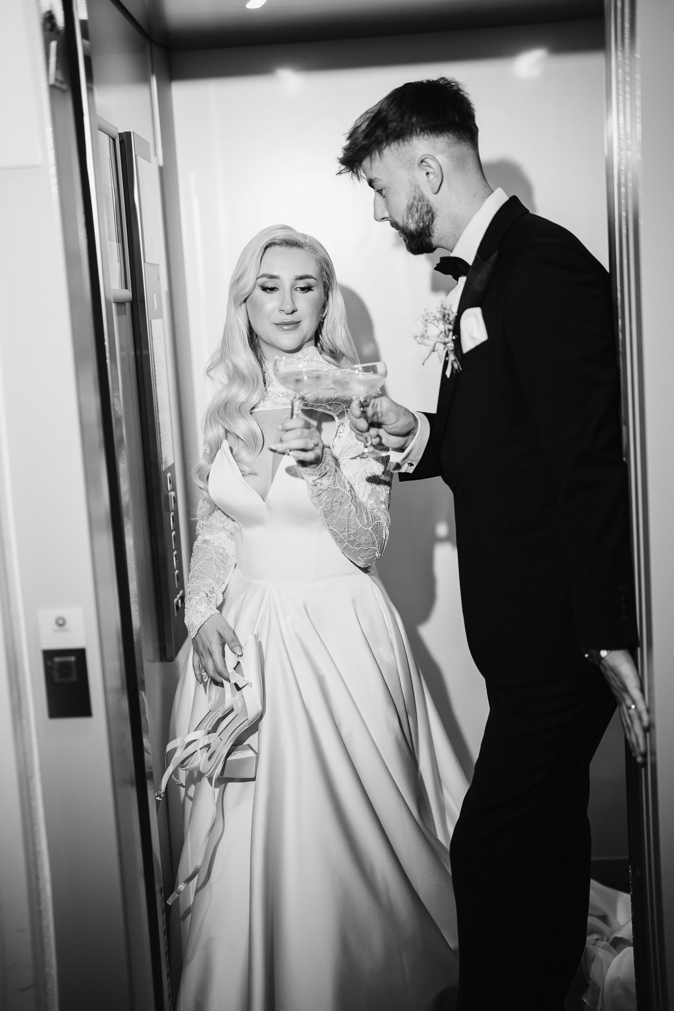 A couple in wedding attire shares drinks in a dramatic black and white corridor setting.