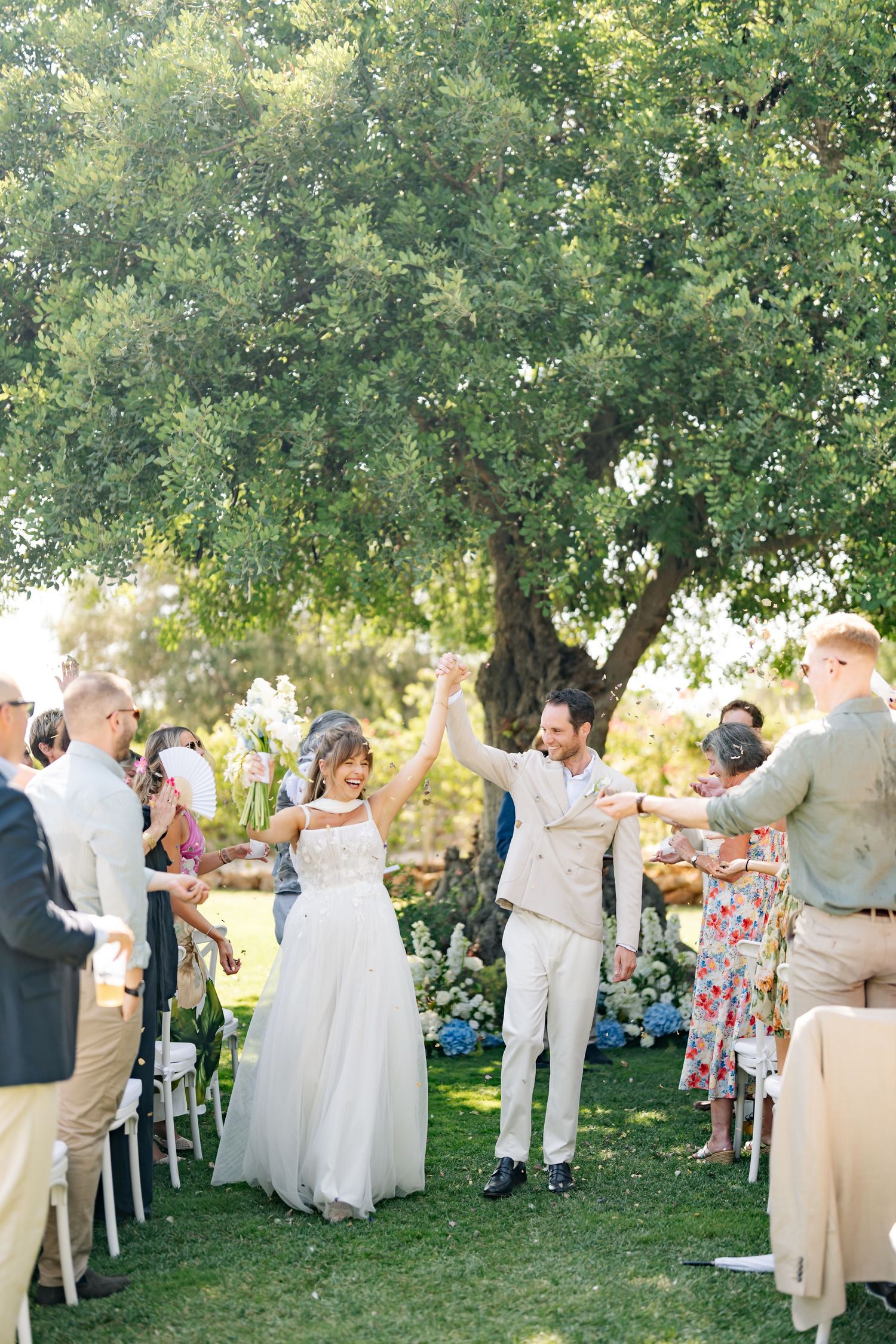 Outdoor garden wedding ceremony with guests seated on white chairs under shady trees during a summer celebration.