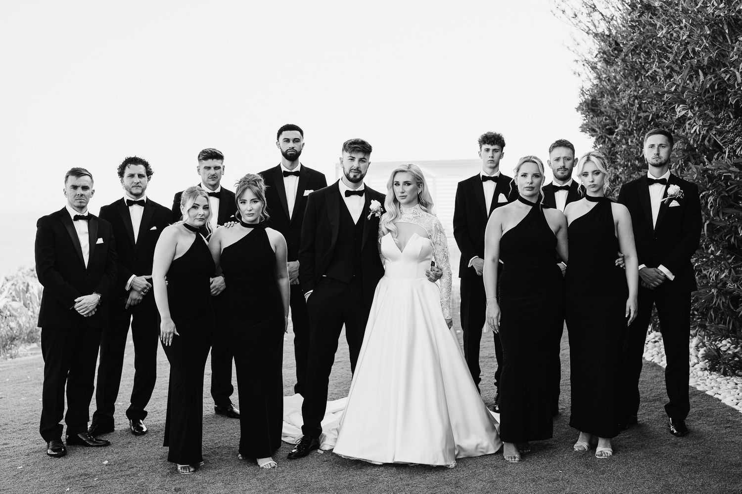 Wedding party members pose together in formal black attire on grassy outdoor venue.