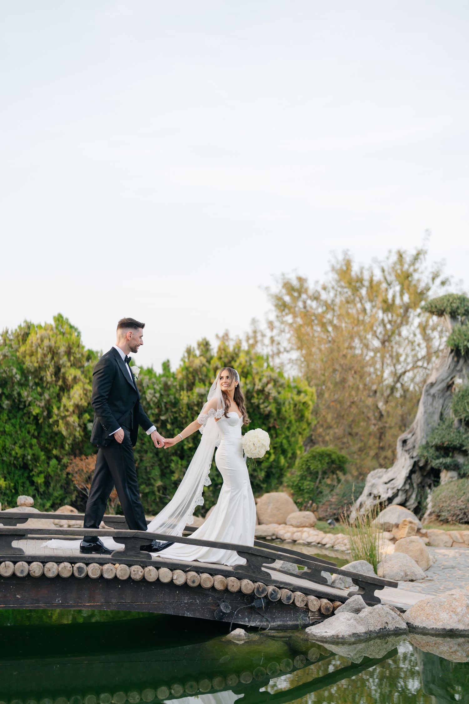 A couple in wedding attire walks across a decorative wooden bridge in a garden setting.