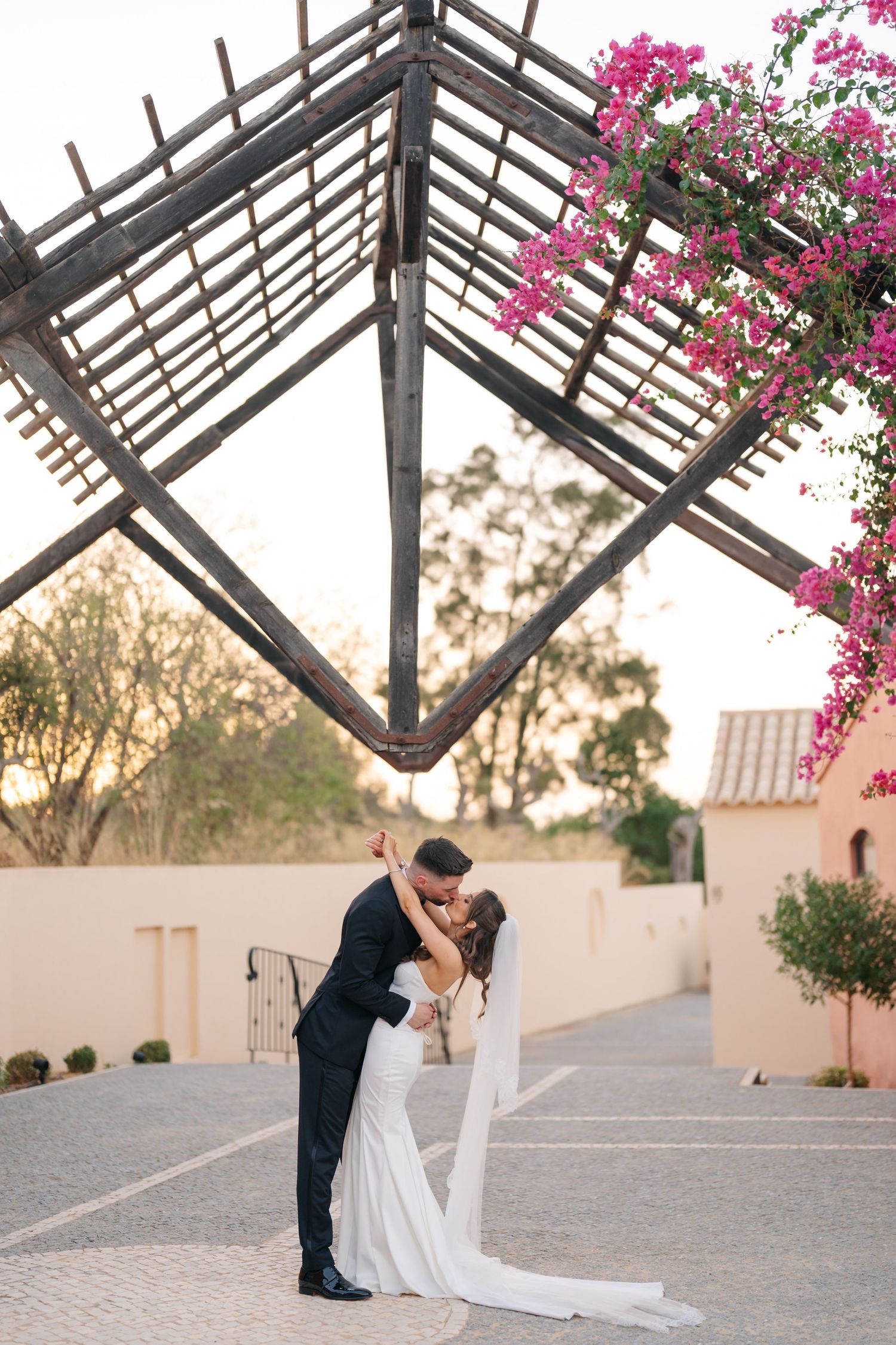 A wedding couple kisses under a wooden pergola with pink bougainvillea flowers.