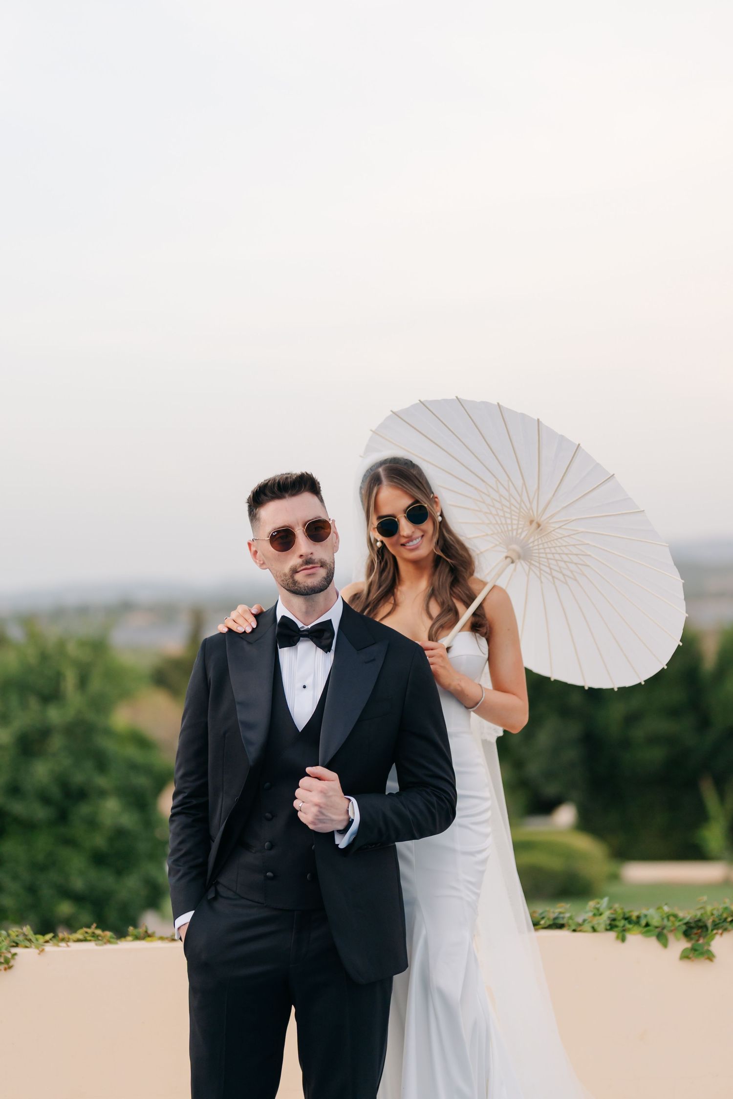 Stylish newlyweds pose with a white parasol against a scenic backdrop at sunset.