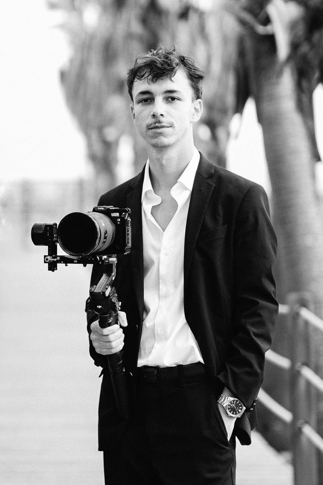 Young man in black suit holding professional camera on gimbal stabilizer outdoors, black and white portrait.