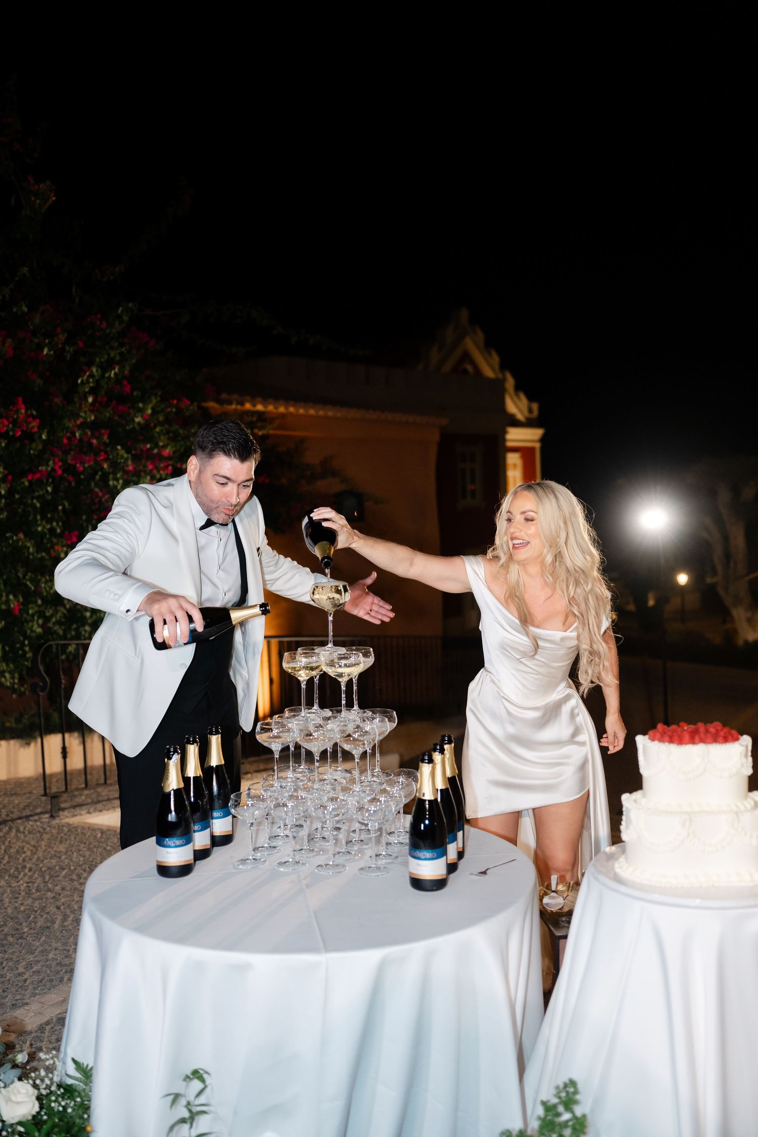 Wedding couple creating a champagne tower with bottles and glasses on a white-clothed table.