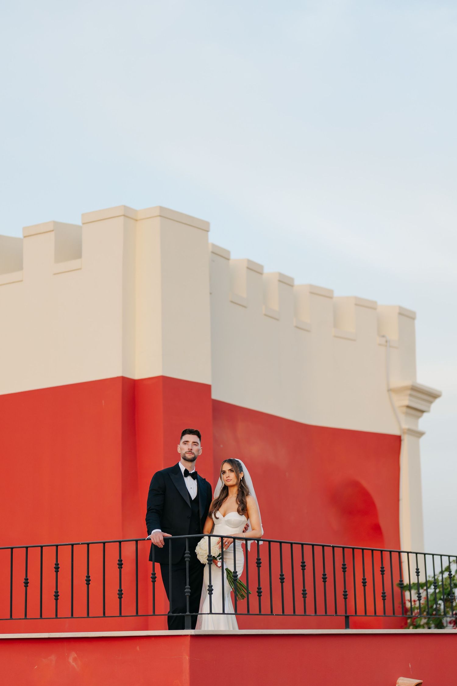 A wedding couple poses on a balcony against a red and white architectural backdrop.