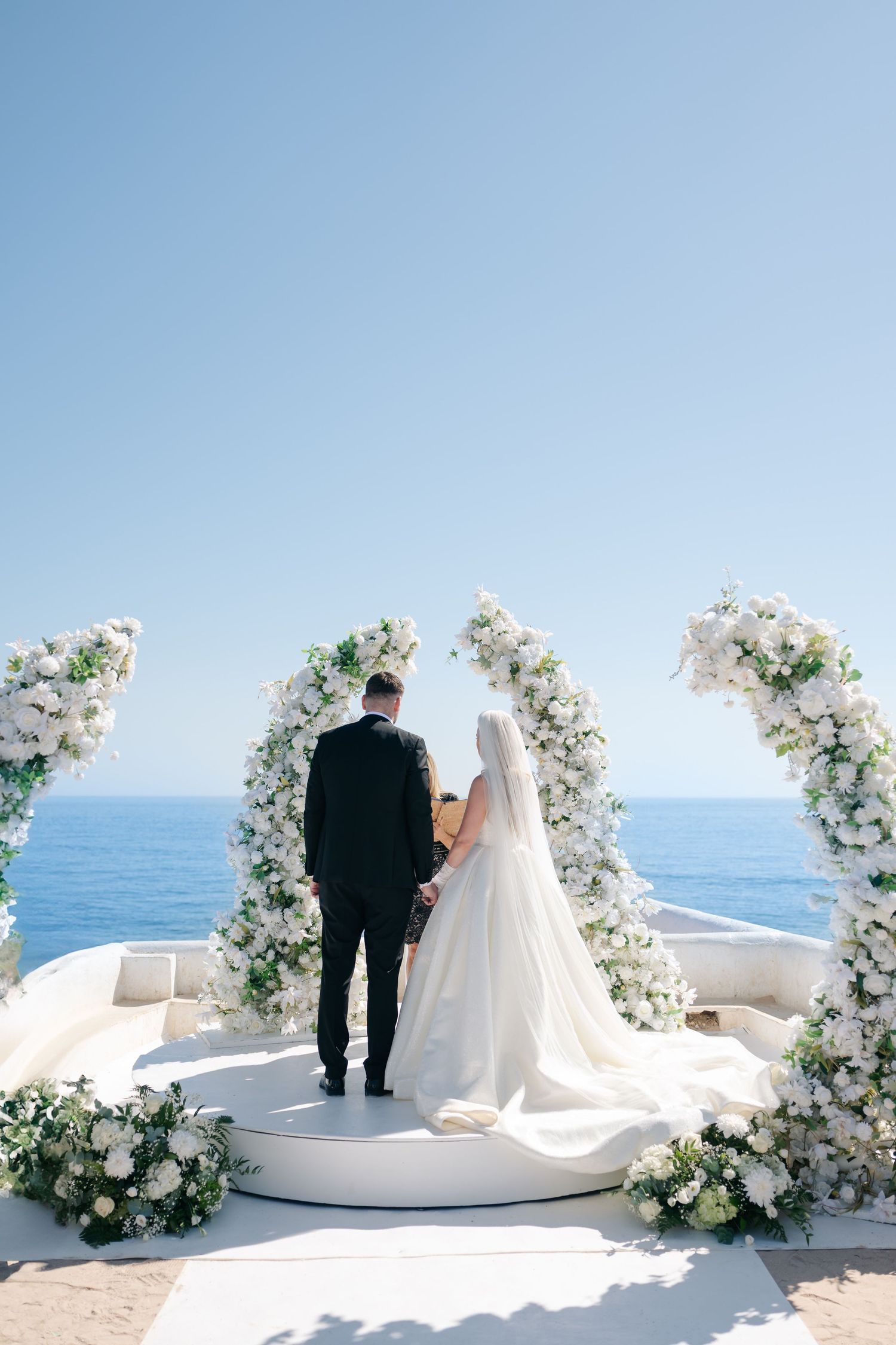 Wedding couple stands under ornate white floral arches on a seaside terrace.