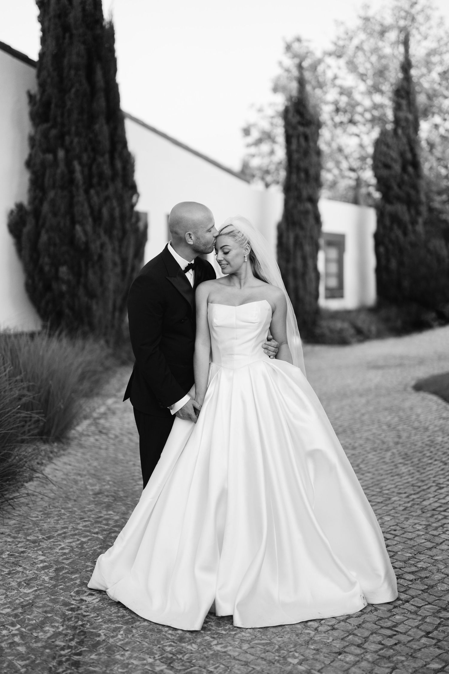 Artistic black and white wedding portrait on cobblestone path with cypress trees.
