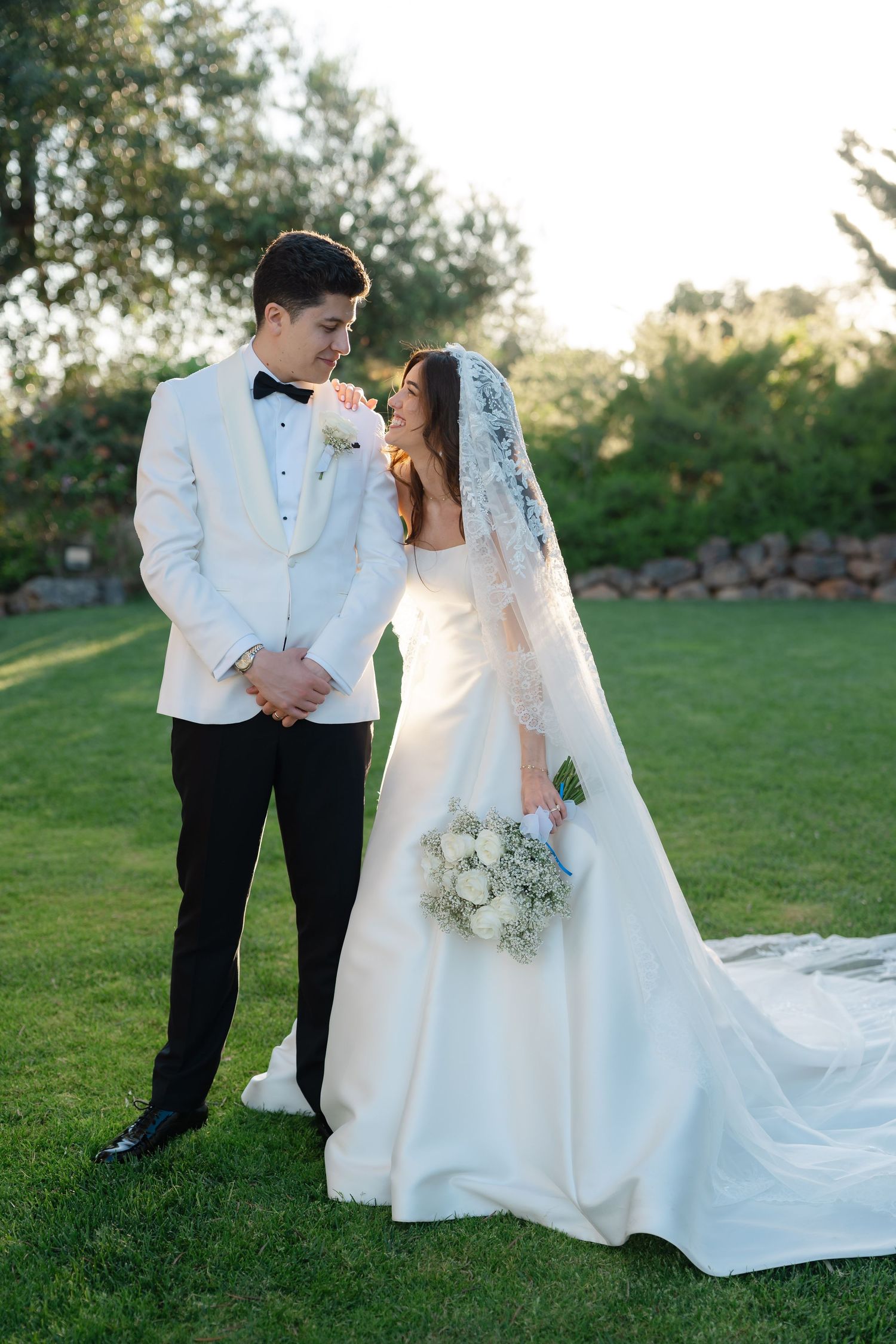 Bride and groom standing on green lawn, she wears a white gown with veil, he wears a white tuxedo jacket.