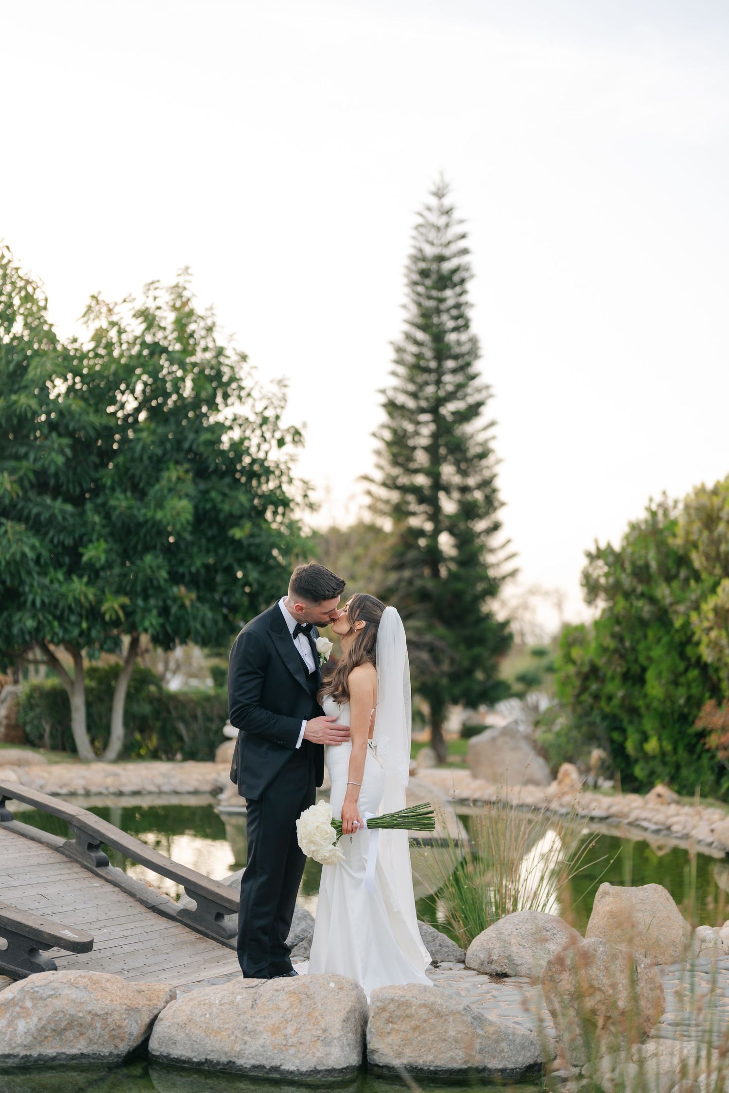 Romantic wedding photo session by a landscaped pond with natural stone features.