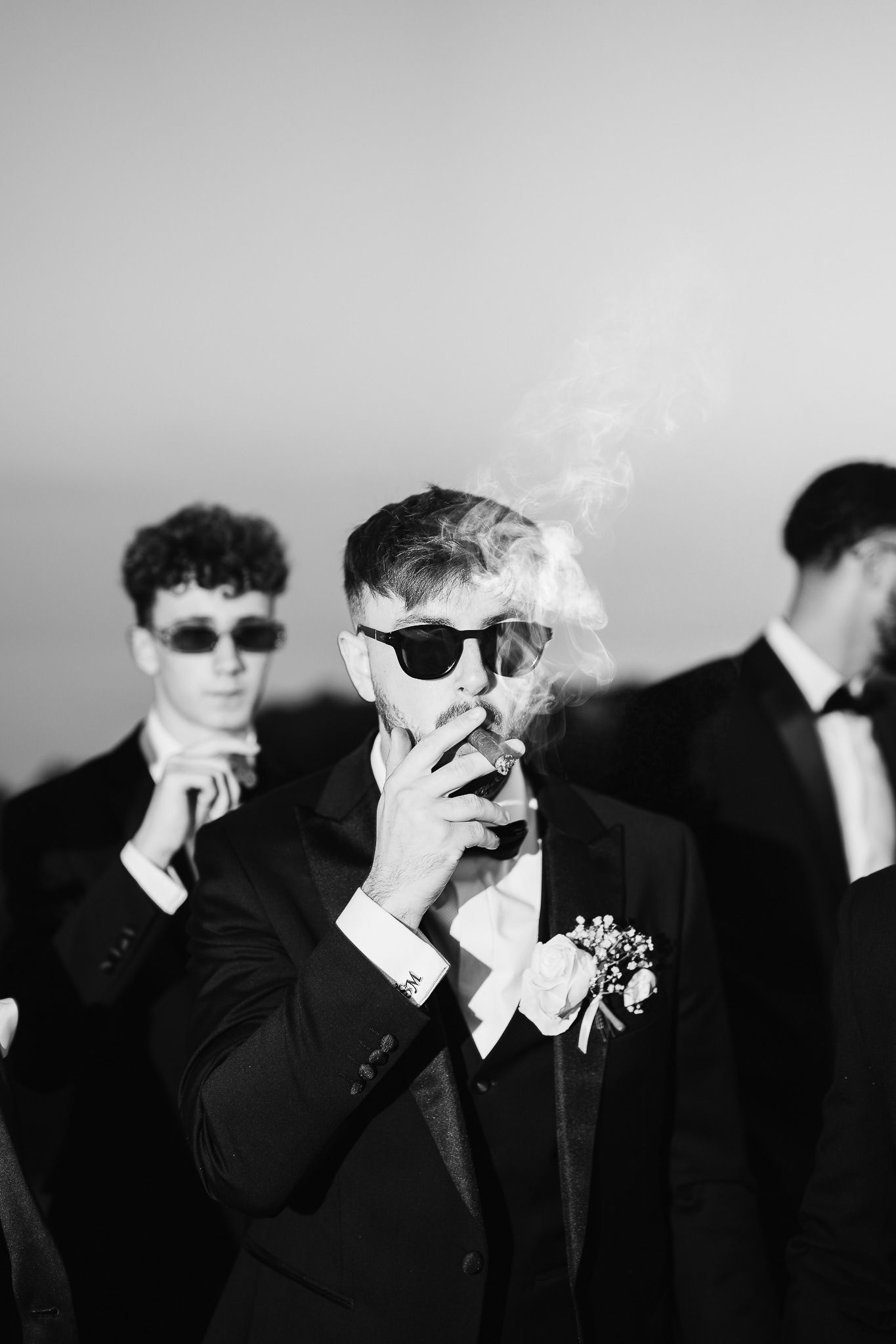 Groomsmen in formal wear light cigarettes while wearing sunglasses in monochrome photography.