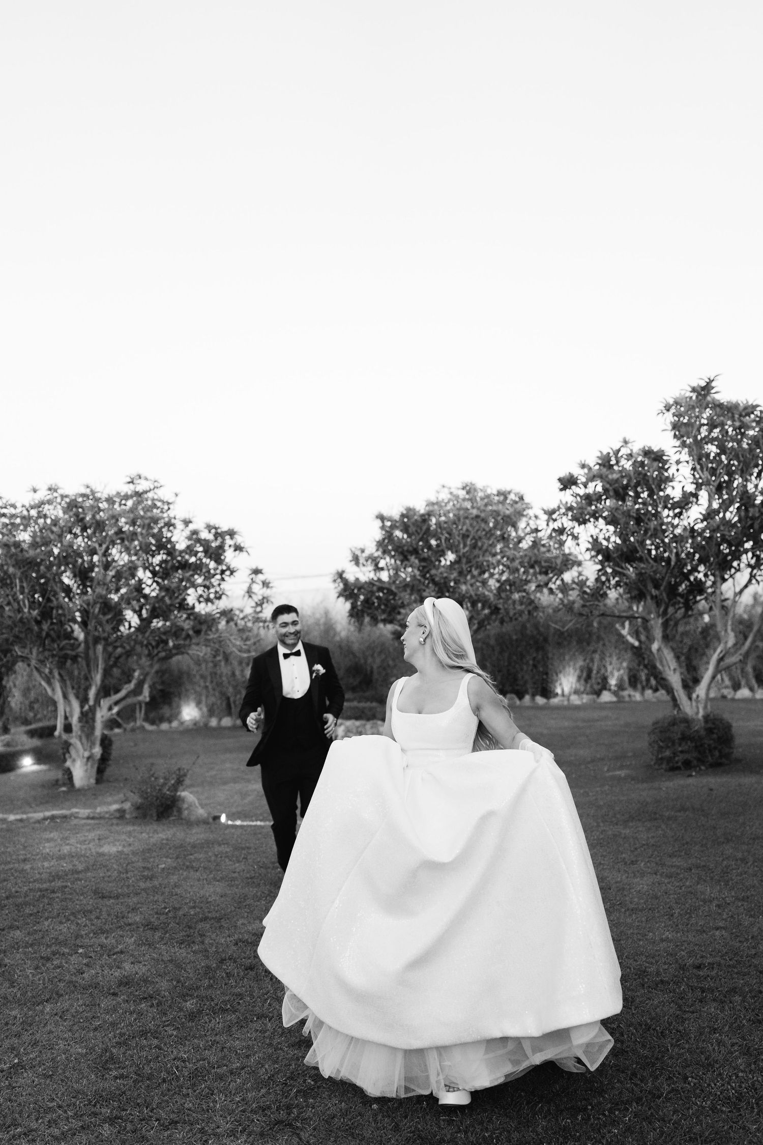 A black and white photo of newlyweds walking together on grass with trees in the background.