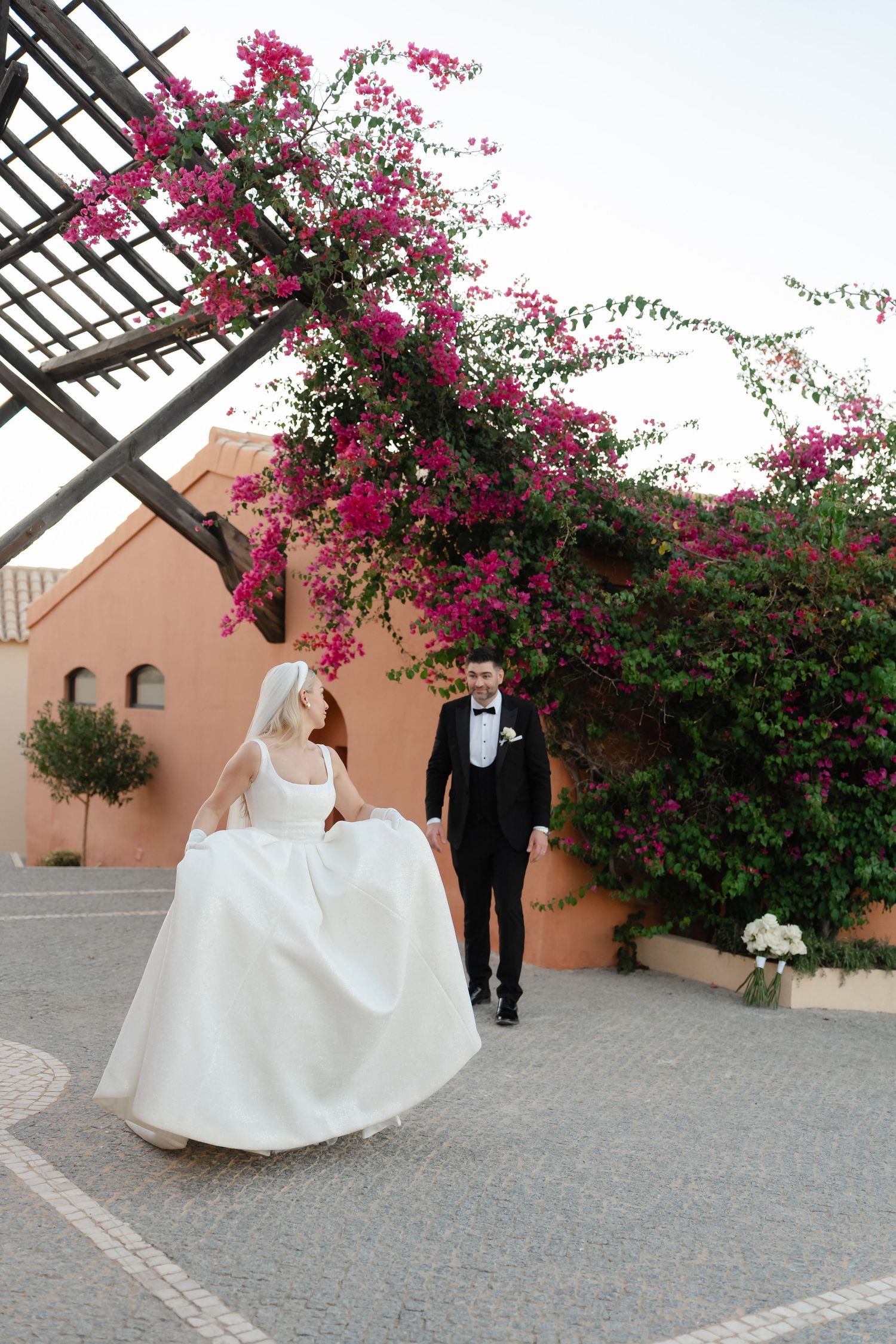 A romantic wedding couple poses under vibrant pink bougainvillea flowers on a paved courtyard.