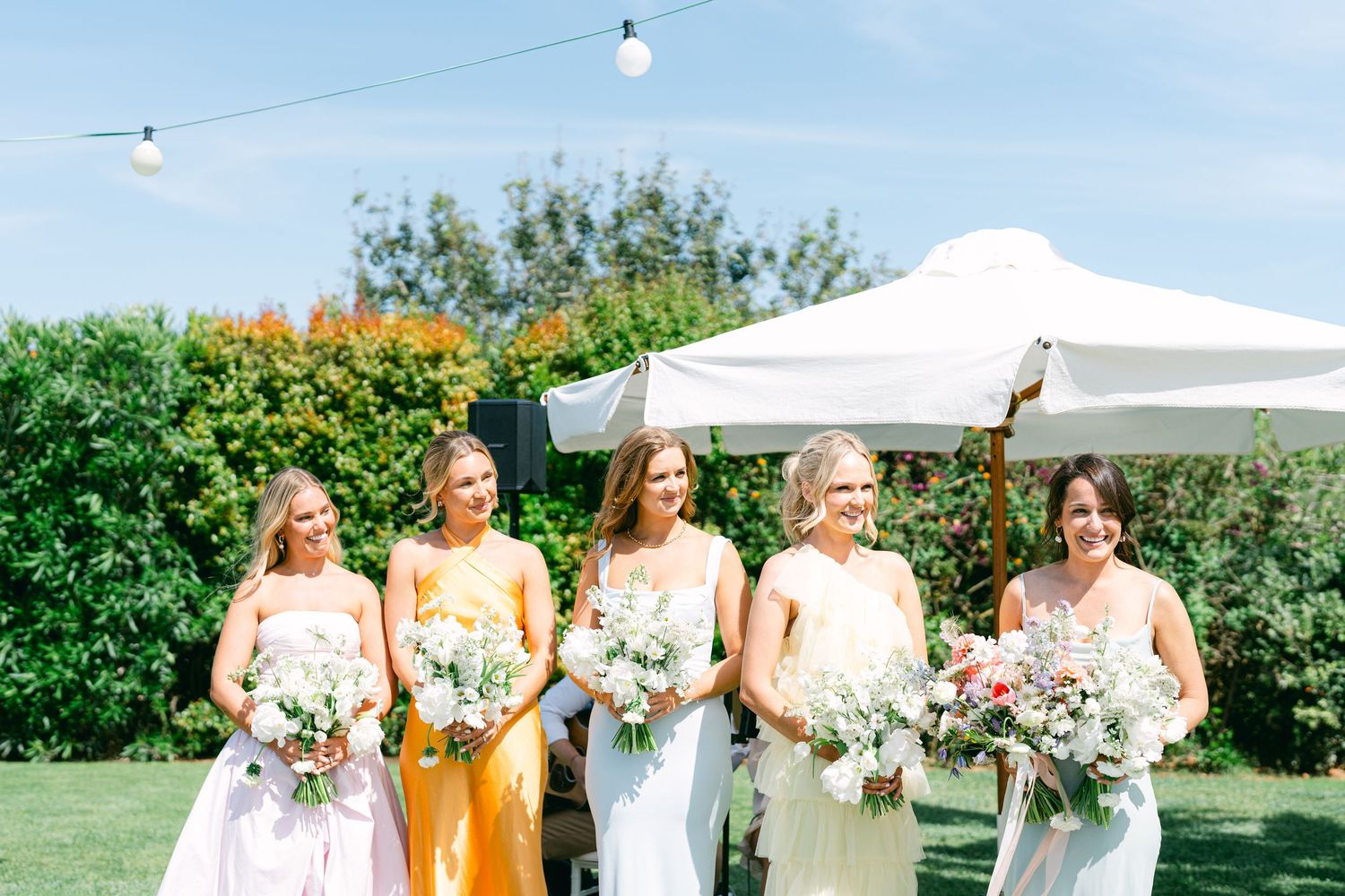 Wedding party in white dresses and yellow accent dress pose together in a tropical garden setting.