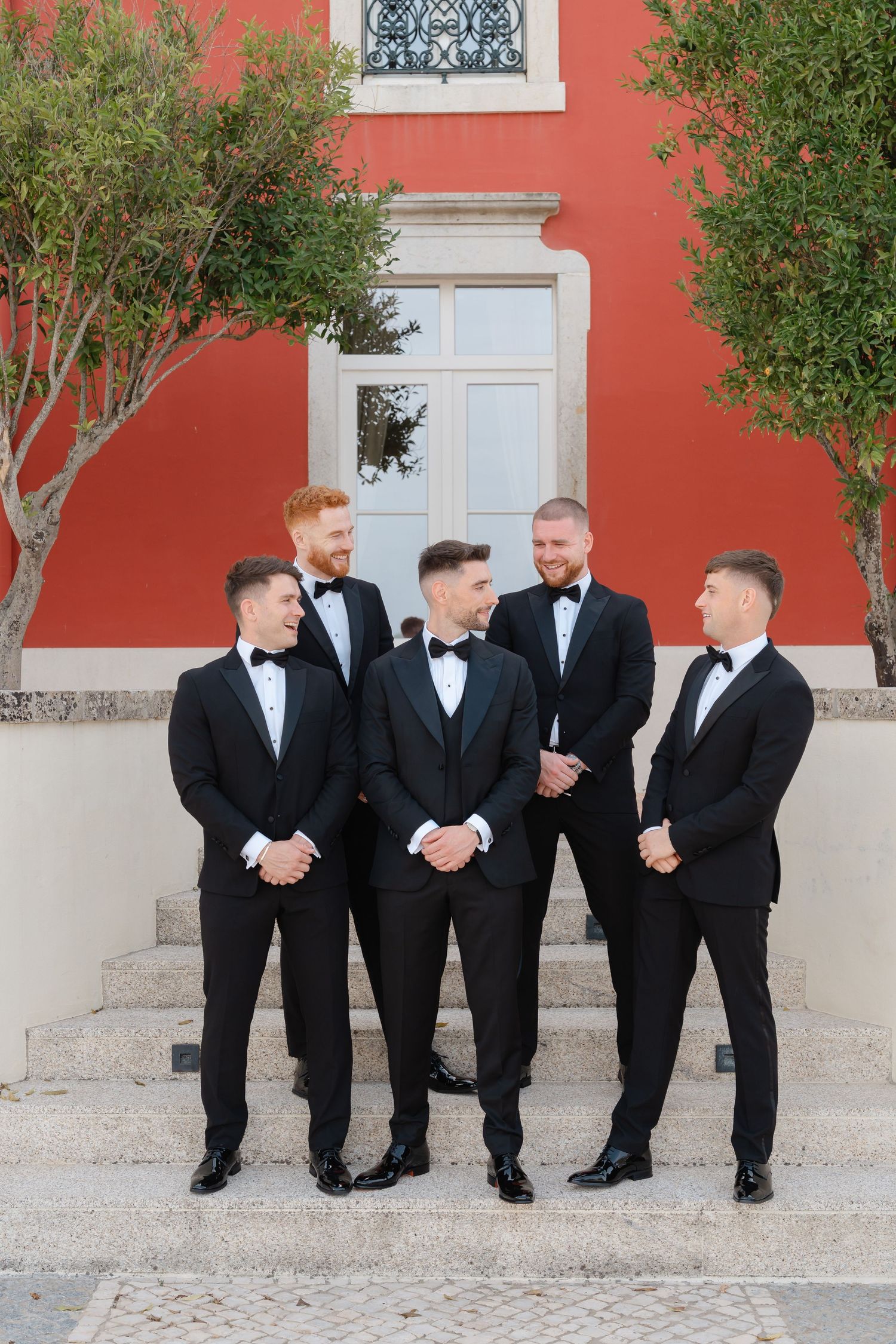 Groomsmen share a candid moment while posing on steps against coral colored building.