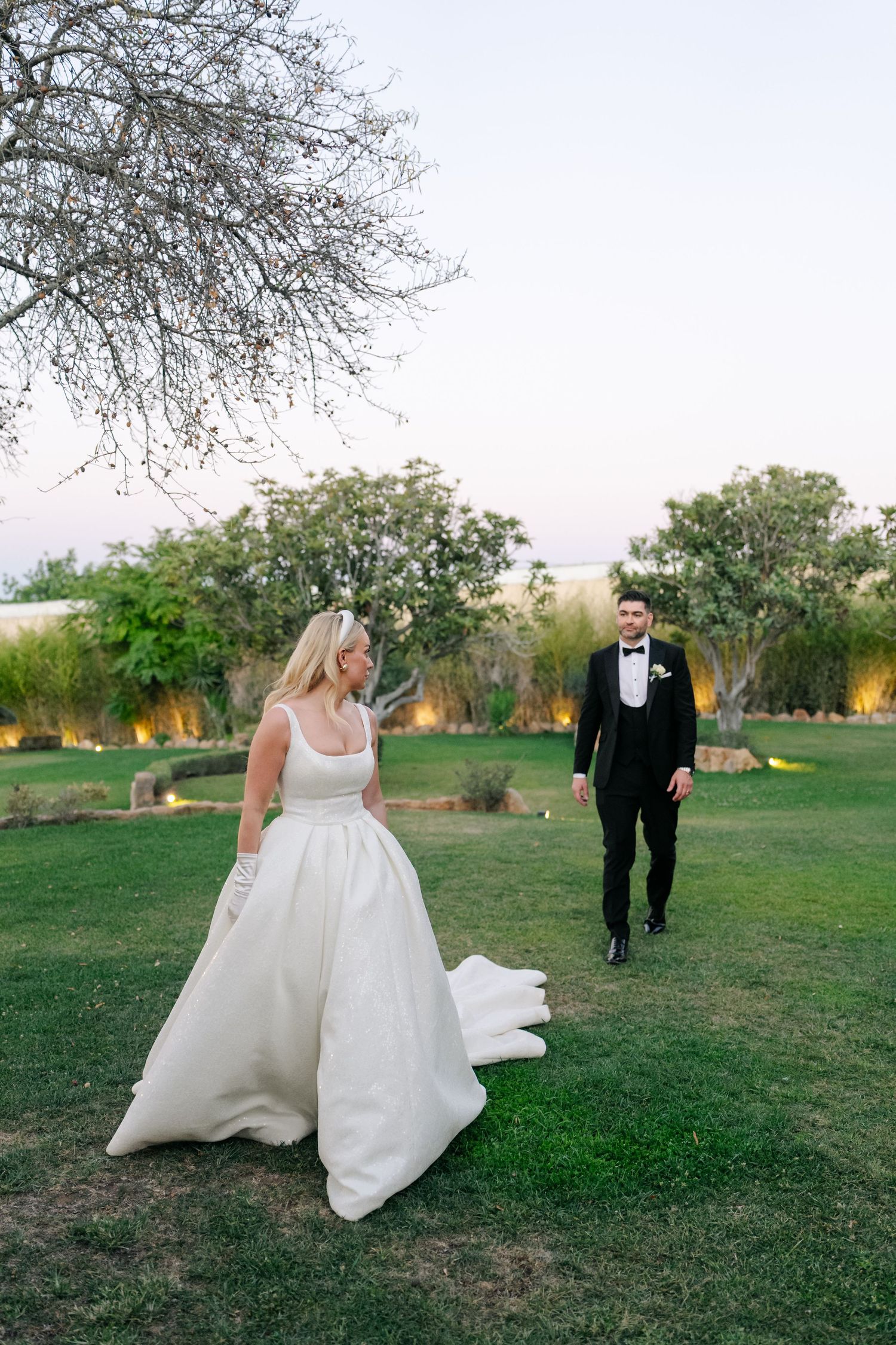 Newlyweds walk together across a grassy lawn with trees in the background during golden hour.