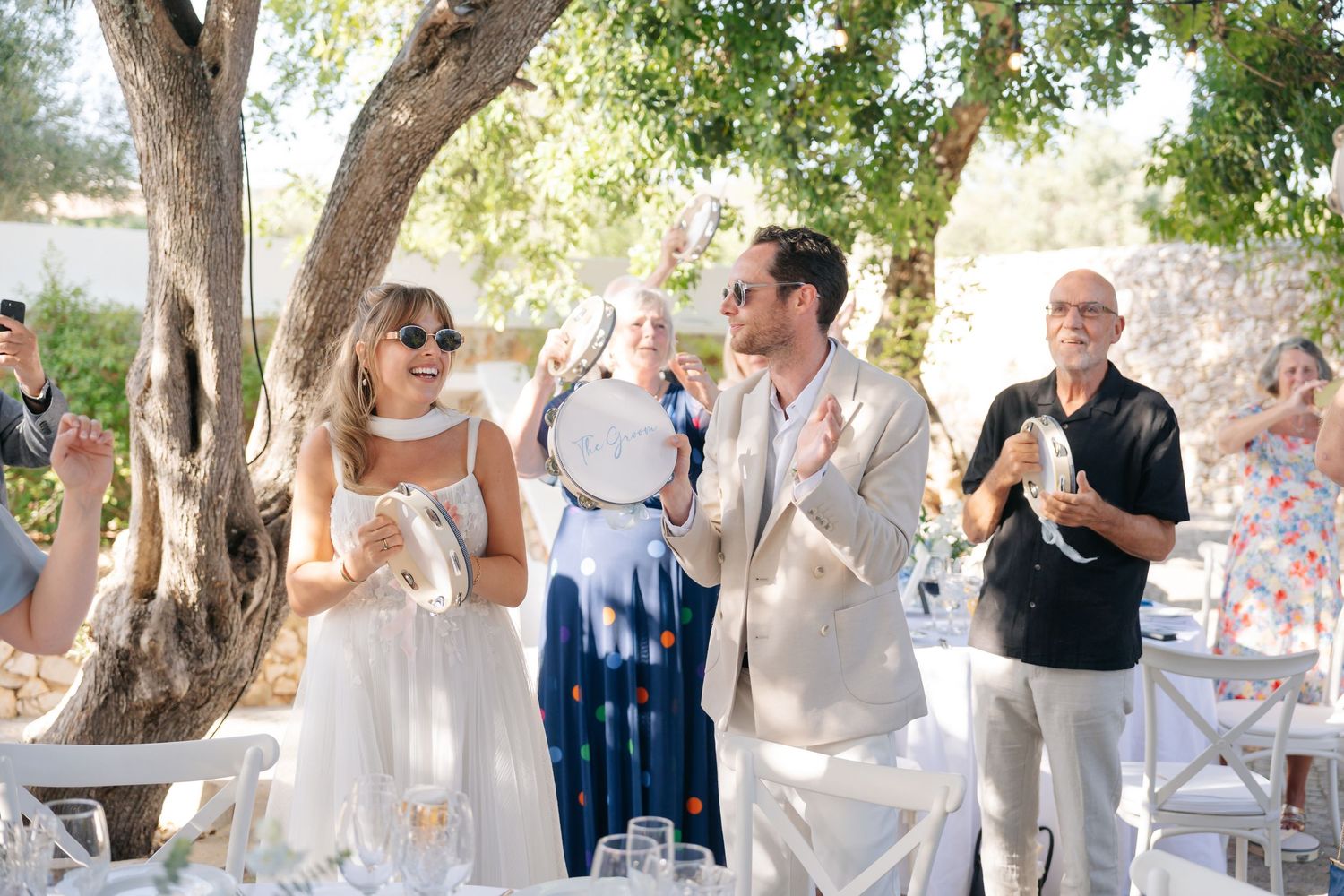 Wedding guests dressed in white gather outdoors under shady trees during a daytime celebration.