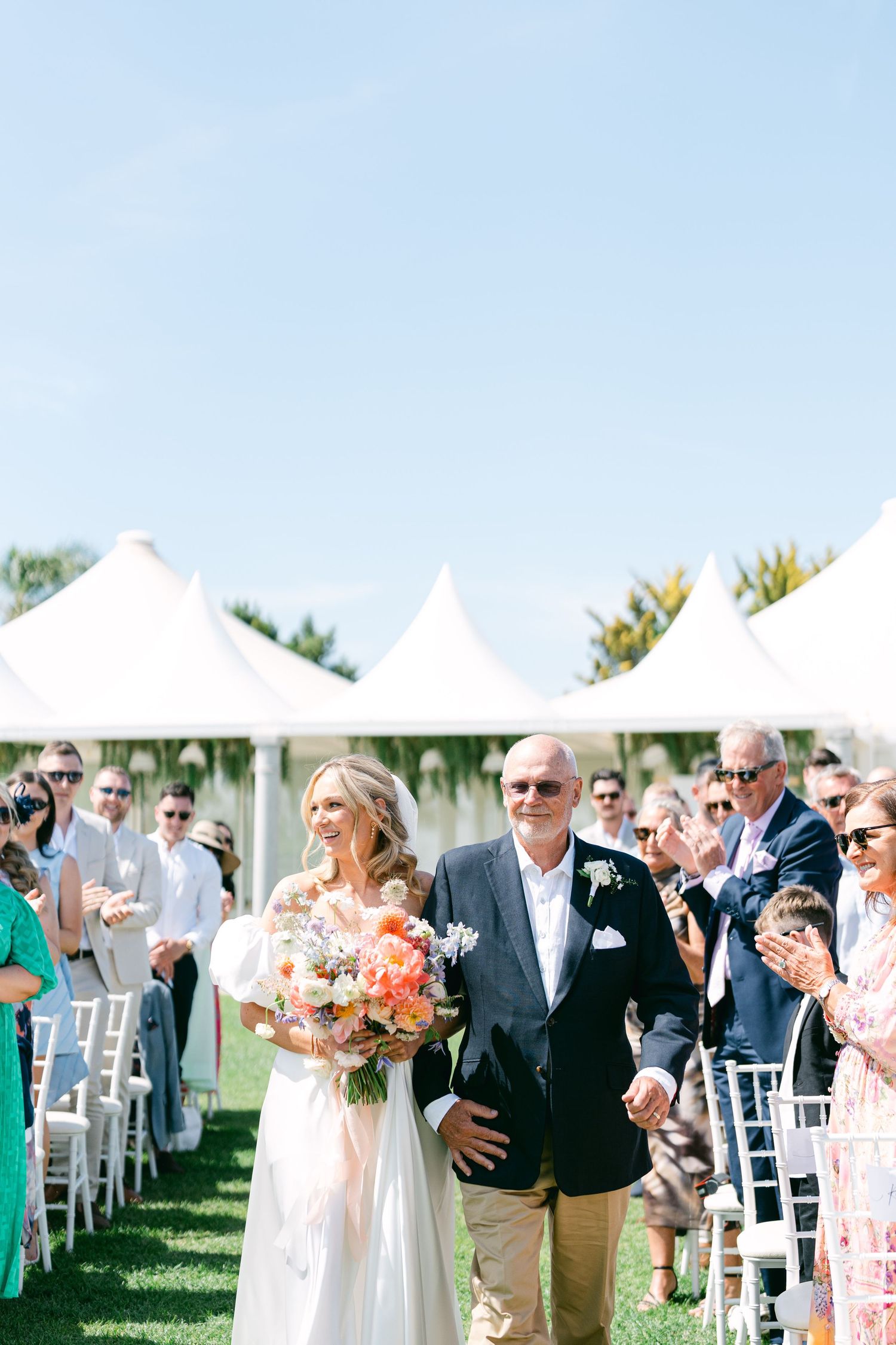 Wedding processional on a lawn with guests seated in white chairs under sunny skies.