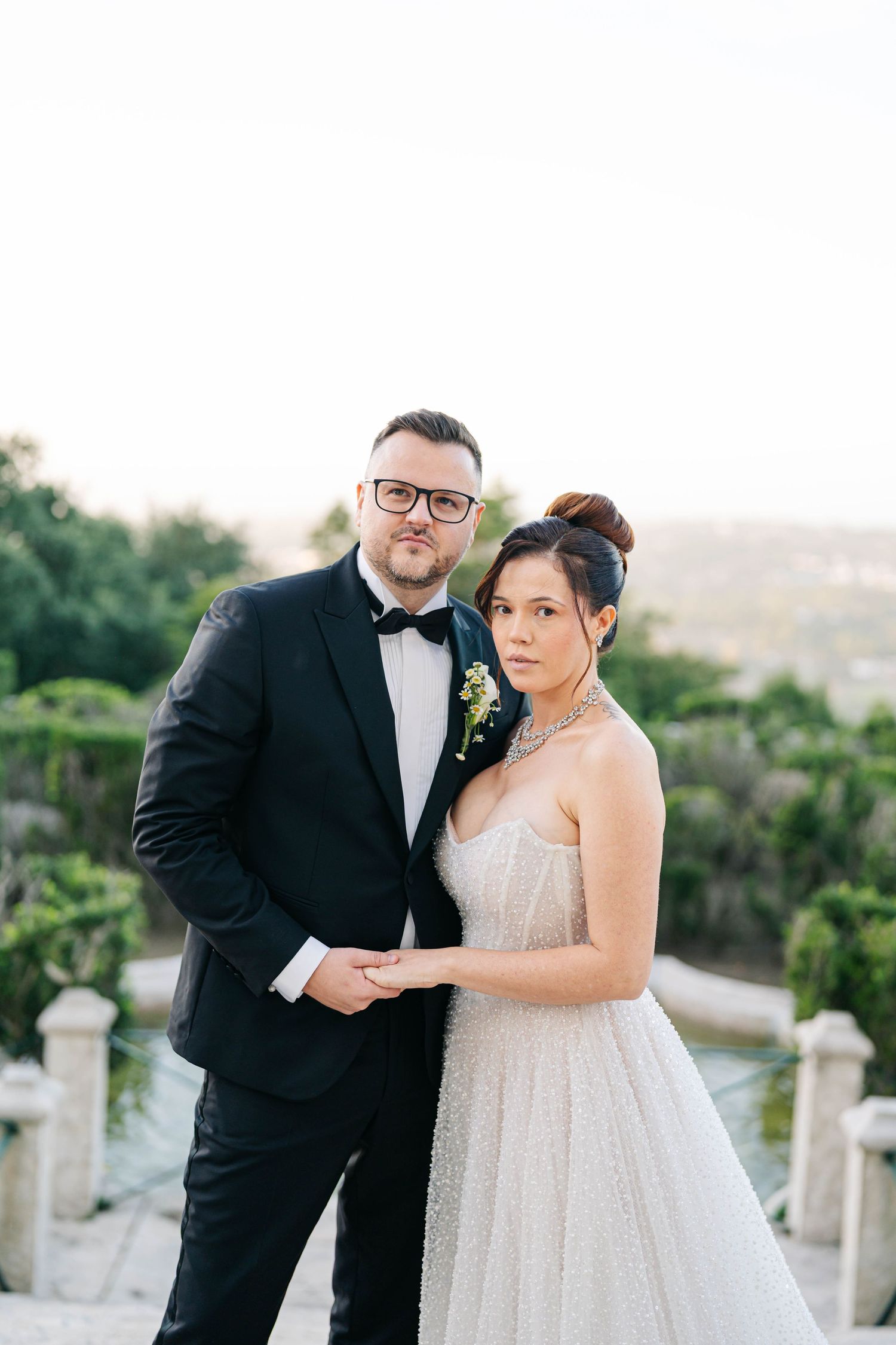 A couple in formal wedding attire poses together outdoors at sunset with lush greenery in the background.