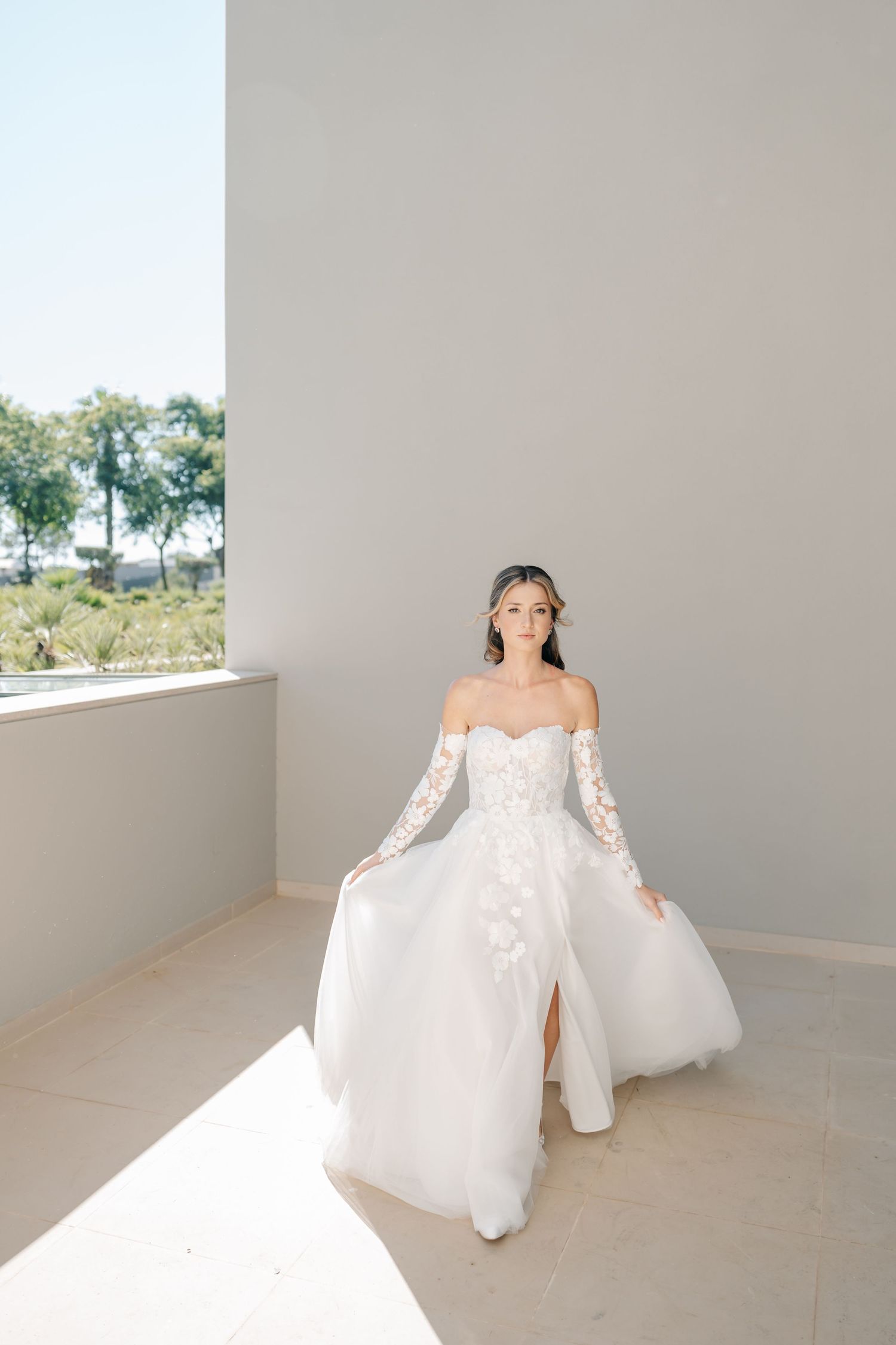 Elegant off-shoulder wedding dress with lace sleeves and thigh-high slit photographed in natural light against neutral wall.