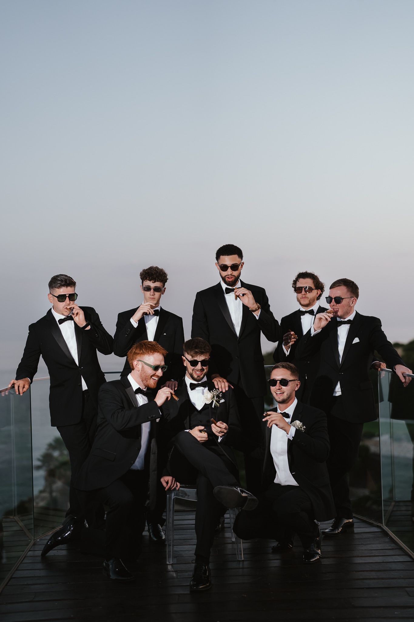 Groomsmen in black suits and sunglasses pose casually outdoors at sunset.