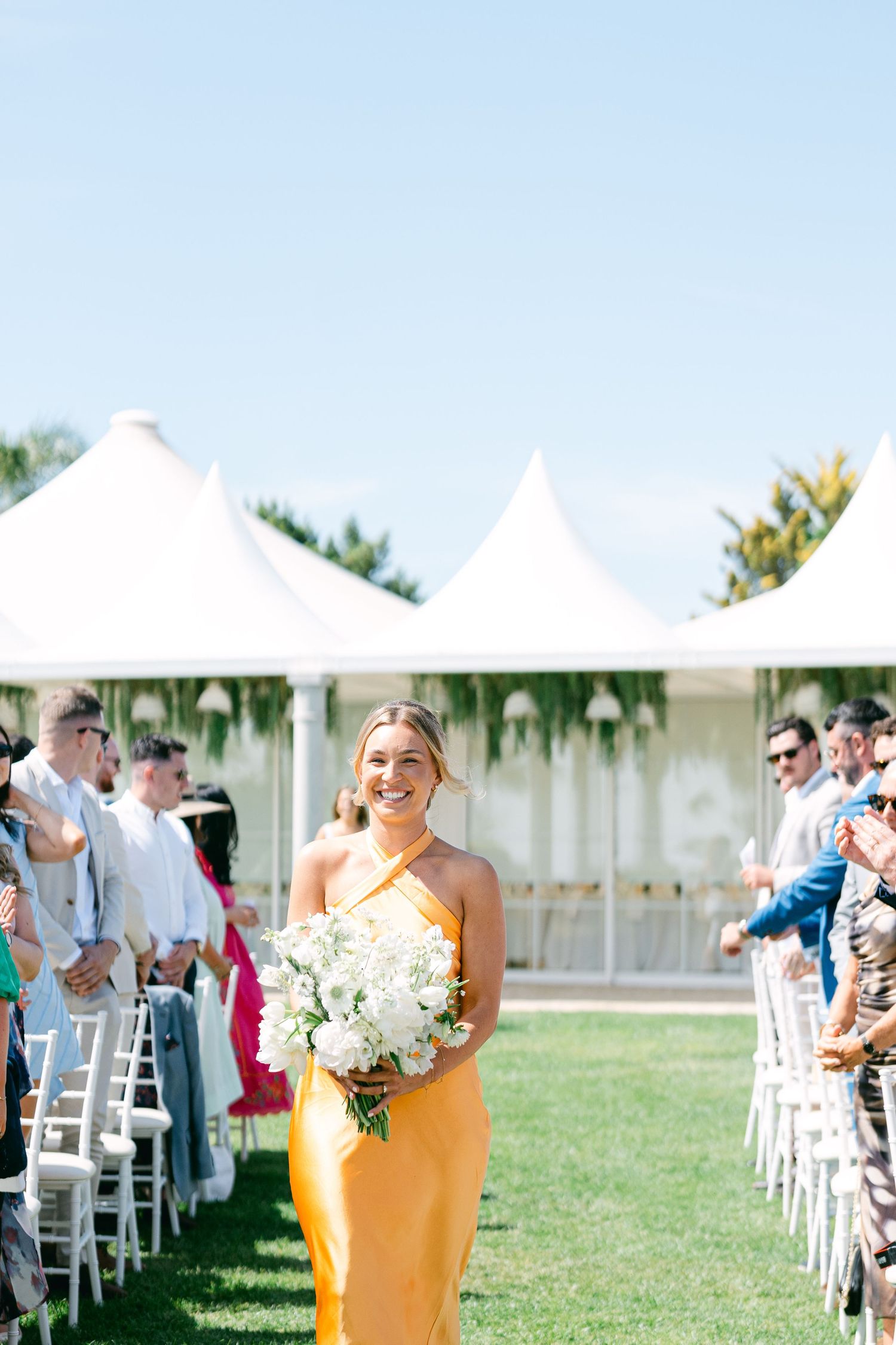 Wedding guests seated in white chairs under white tents during an outdoor ceremony.